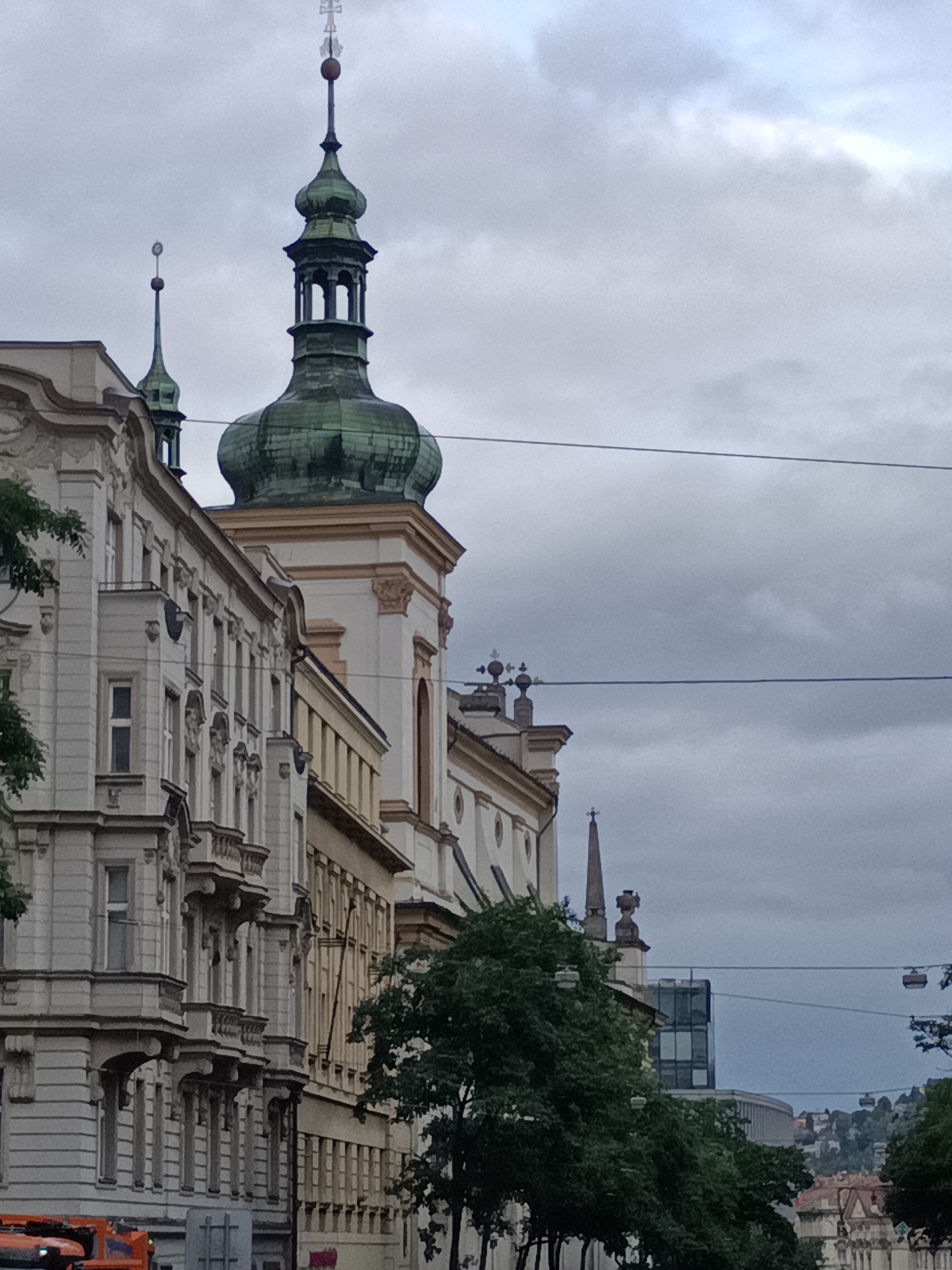 The image shows a view of St. Nicholas Church in Malá Strana, Prague, Czechia. The church's Baroque-style architecture is visible, with its prominent green dome and spires. The sky is overcast, and there are some trees in the foreground. The image is taken from street level, looking up at the church.