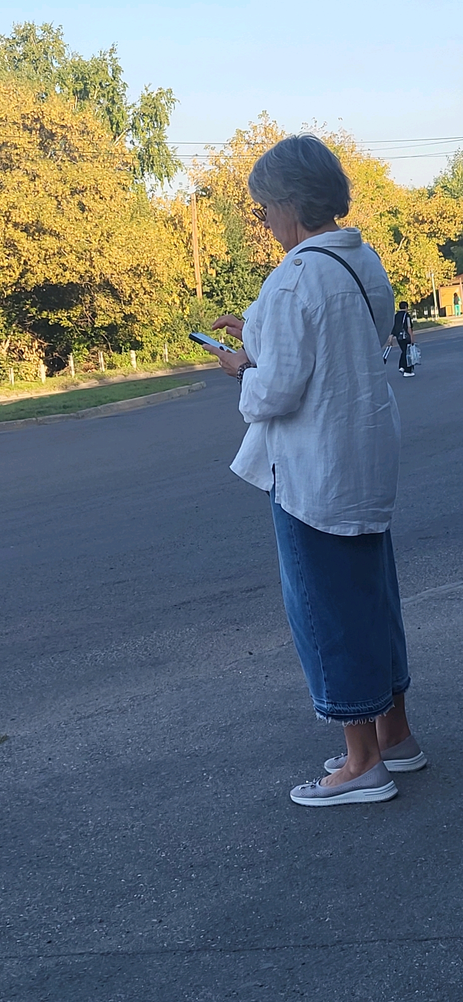 The image shows a woman standing on a paved road, looking at her phone. She has gray hair and is wearing a white linen shirt and a long denim skirt. She is also wearing grey slip-on shoes. The background shows trees with yellowing leaves, suggesting it is autumn. There is another person in the distance walking with a bag. The scene appears to be on a street in Oskemen, Kazakhstan, given the context of the request.