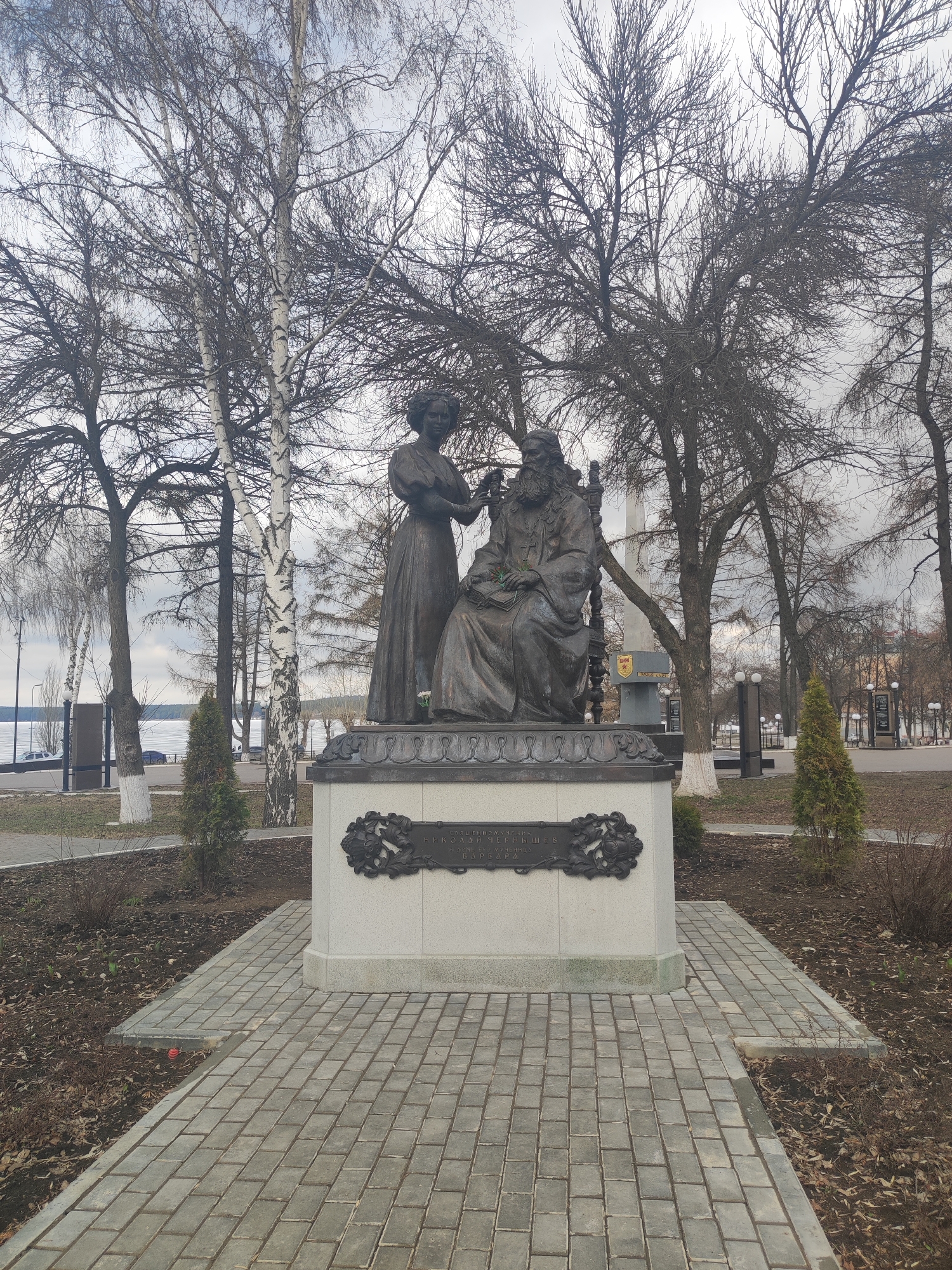 The image depicts a bronze statue of Nikolai Chernyshevsky, a Russian revolutionary democrat, philosopher, critic, and socialist, located in the city of Votkinsk, Russia. Chernyshevsky is seated in a chair, holding a book. A woman stands beside him, seemingly placing a wreath on his head. The statue is situated on a light grey stone pedestal with a dark metal plaque bearing inscription in Russian. The pedestal sits on a platform of grey brick pavers. Behind the statue, there are several bare trees, including birch trees. In the background, there are additional trees, street lights, and bodies of water are visible. The sky is overcast, suggesting a cloudy day. The statue is surrounded by a garden area with bare earth and small evergreen shrubs.