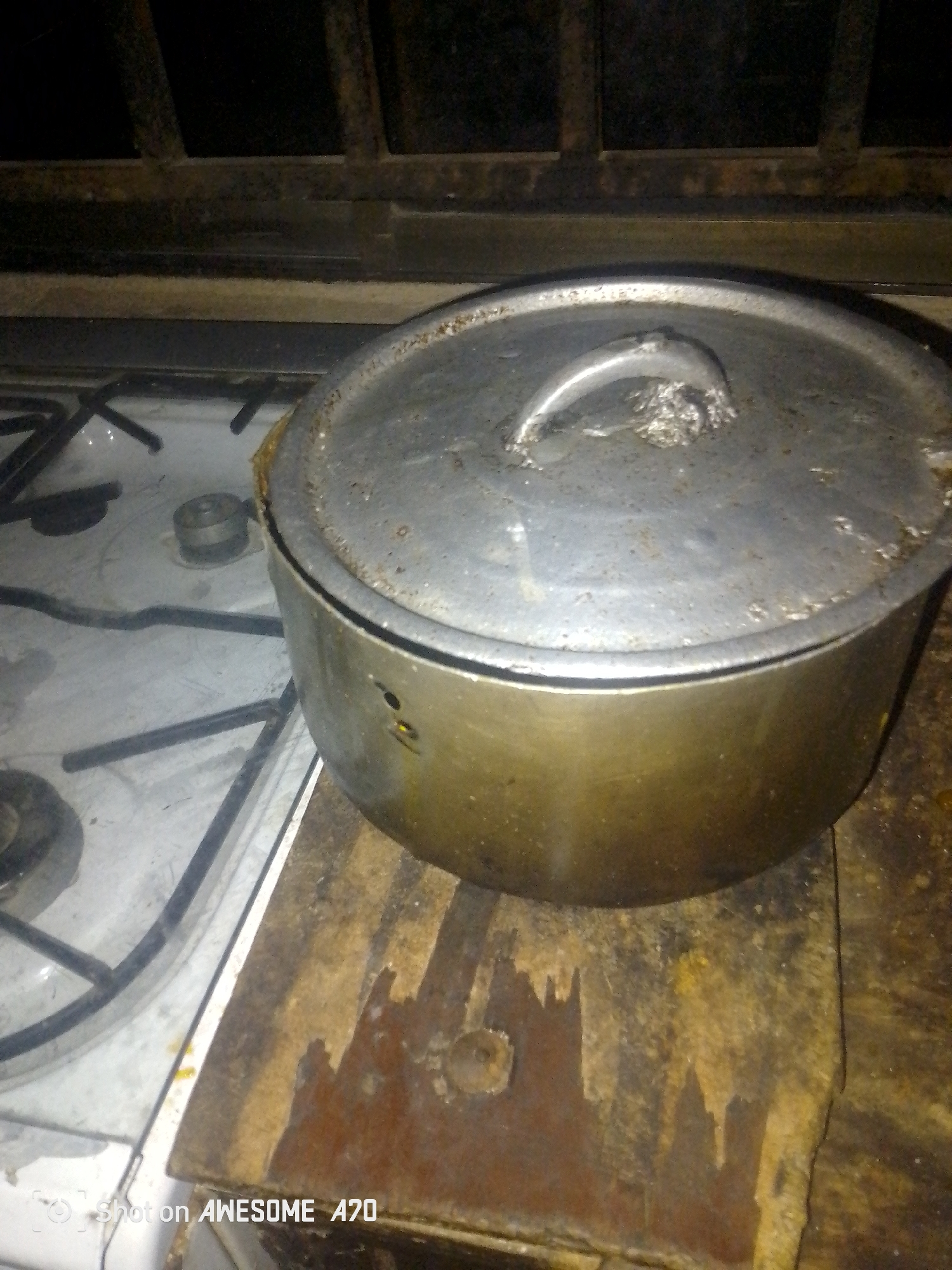 A silver pot with a lid is placed on a wooden surface next to a gas stove. The pot is round and appears to be made of aluminum. The lid has a handle that is attached to the top. The gas stove has four burners and a white surface. The wooden surface is old and appears to be stained and worn. The background is dark and out of focus, with a suggestion of a metal grate. There are no people or animals visible in the image. The lighting is dim, creating shadows and highlighting the textures of the objects. The image is taken from a slightly high angle, looking down at the pot and stove. The timestamp "Shot on AWESOME A70" is visible in the bottom left corner.