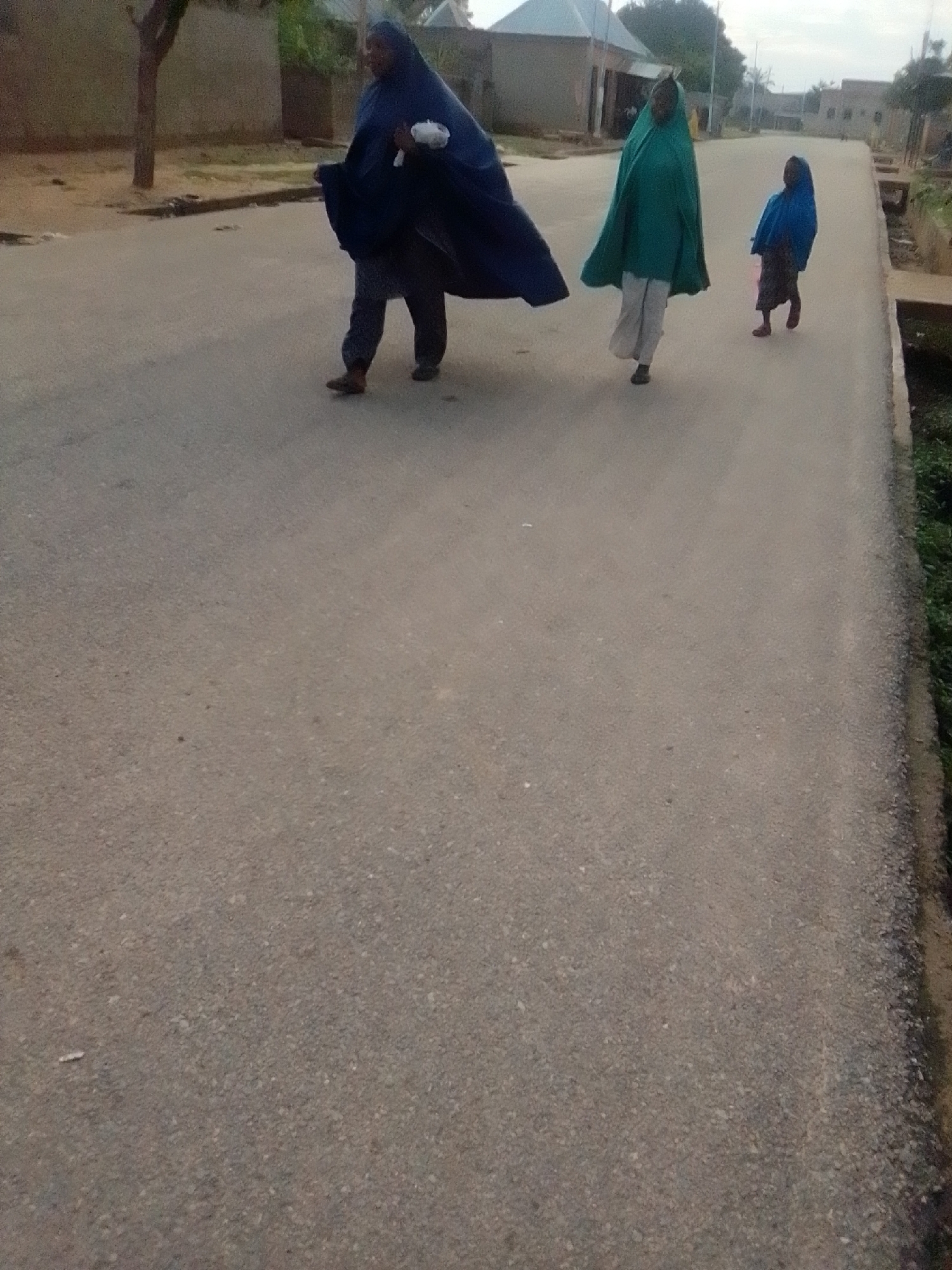 Three individuals are walking on a paved road in what appears to be a residential area in Liji Hill, Nigeria. The person in the foreground on the left is wearing a dark blue hijab and a darker garment, carrying a white bundle in their left hand. This individual is also wearing trousers and brown shoes. To their right, a second person is wearing a green hijab and light-colored trousers, walking away from the camera. Further to the right and slightly behind, a third person, likely a child, is wearing a blue hijab and a dark skirt, also walking away from the camera. The road surface is asphalt, with some scattered debris. The surroundings include buildings with light-colored walls and corrugated metal roofs, and vegetation along the side of the road. The lighting suggests it is daytime, possibly late afternoon or early morning due to the diffused light.