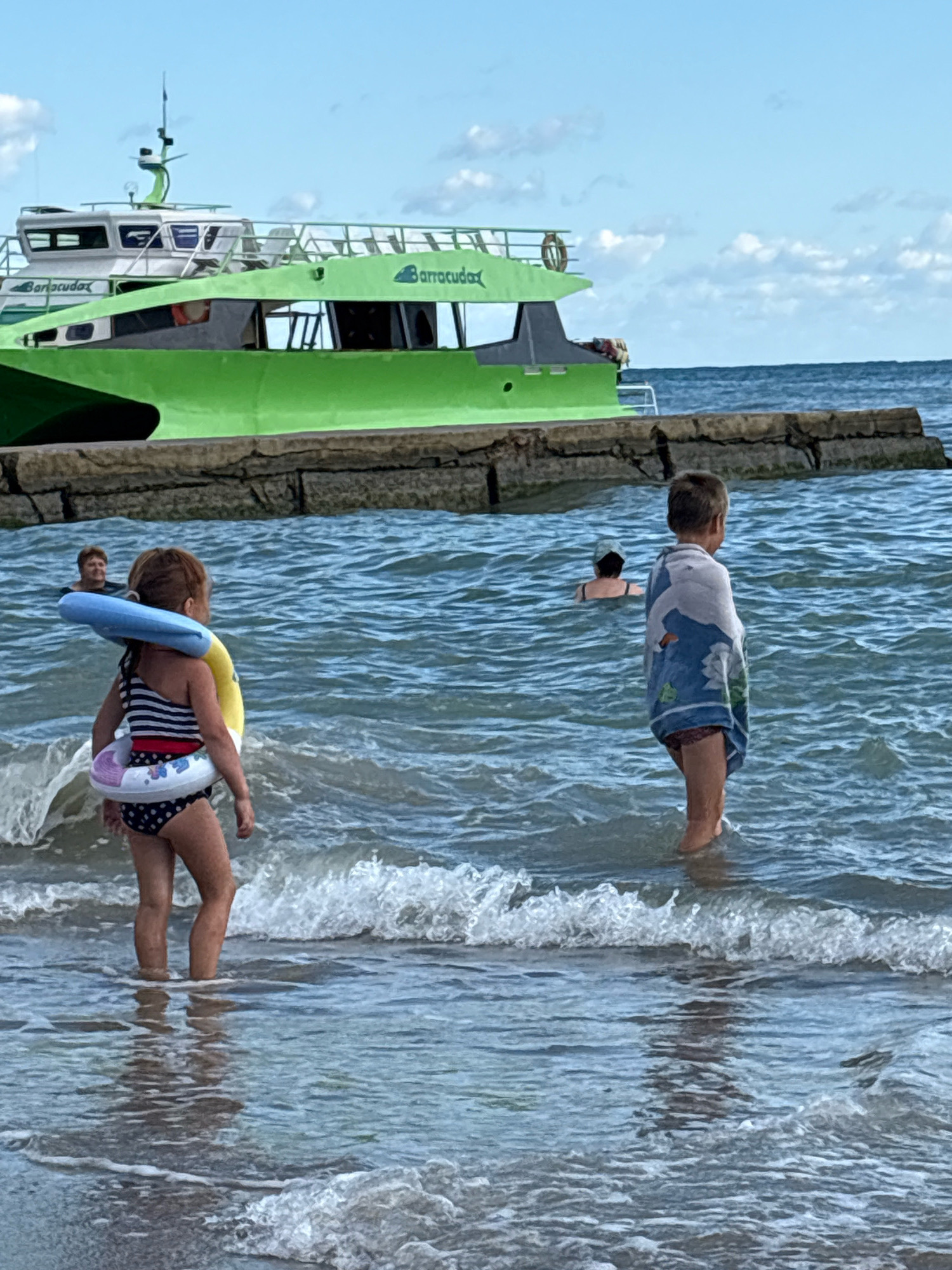 This image captures a bright, daytime scene at a coastal area, likely in Arkhipo-Osipovka, Russia, given the context. In the foreground, a young girl wearing a striped and polka-dot swimsuit stands in shallow water, with a blue inflatable ring around her neck and a smaller inflatable ring around her waist adorned with floral patterns. Her legs are partially submerged, and small waves are breaking around her. To her right, a boy, seen from the back, stands in the water wearing a towel with a blue and white dolphin pattern wrapped around him. Further in the background, a person is partially visible in the water, with their head and shoulders above the surface. The water is a deep blue with visible ripples and waves, suggesting it is the sea. Beyond the water, a grey stone pier or breakwater runs horizontally across the midground. Behind the pier, a vibrant green boat with a white cabin is docked or anchored. The boat has "Barracuda" written on its side in green lettering. The sky above is blue with scattered white clouds. The overall scene depicts a relaxed, recreational setting by the sea, with children enjoying the water and a boat present.