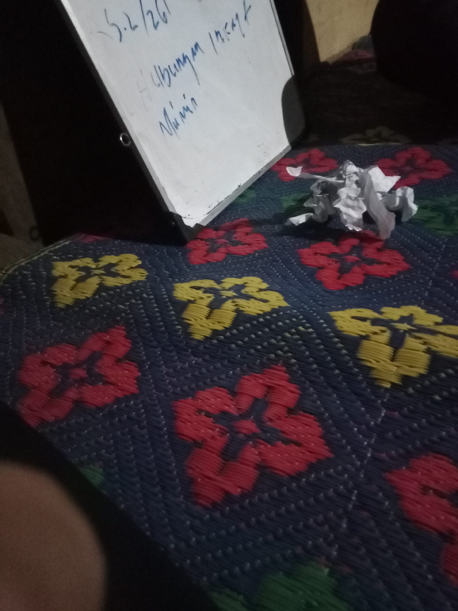 The image captures a whiteboard with handwritten notes and a crumpled piece of paper on a patterned mat. The whiteboard, tilted and angled to the left, displays text in blue ink, including "5.2/26" and "Hubungan in..." which is partially obscured. Below this, the word "Mumin" is clearly visible. The crumpled paper lies to the right of the whiteboard, appearing to be white. The mat beneath is dark blue with a woven pattern, featuring repeating floral designs in red and yellow. The scene is dimly lit, with shadows cast on the wall behind the whiteboard and a soft focus effect. The overall impression is one of a casual, indoor setting, possibly a desk or table area. There is no discernible location in Liwa, Indonesia that can be identified from the visual information provided.