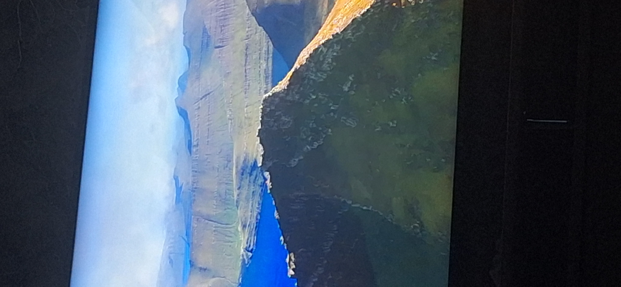 The image displays a breathtaking aerial view of a rugged, mountainous landscape meeting a tranquil blue body of water. The foreground is dominated by a steep, grassy mountain slope with rocky outcrops. Beyond this, layers of terraced mountains recede into the distance, their slopes etched with visible lines, suggesting agricultural or natural formations. A vibrant blue sea or large lake flanks the coastline. The sky above is a clear, pale blue, transitioning into lighter hues near the horizon. The image does not depict the city of Bila Tserkva, Ukraine.
