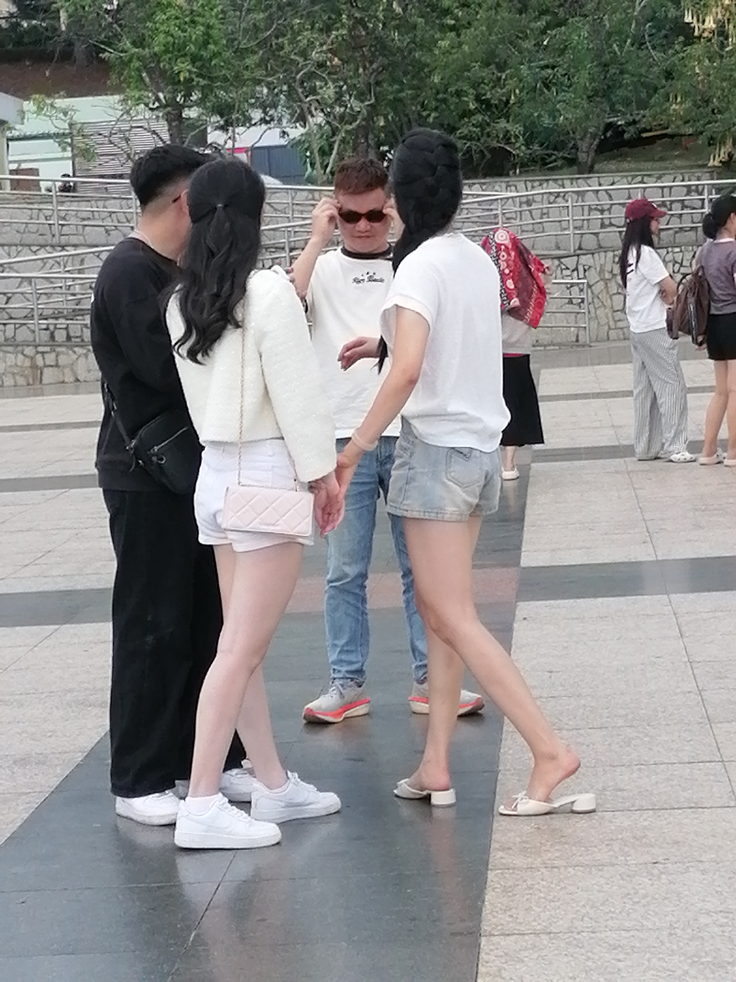 In Da Lat, Vietnam, four individuals are gathered on a paved outdoor area with a stone wall and greenery in the background. A man on the left, wearing a black long-sleeved shirt and black pants, has a black crossbody bag. To his right, a woman with long black hair tied in a ponytail is wearing a white textured jacket over a white top and white shorts, carrying a light pink quilted purse. In the center, a man in a white t-shirt and blue jeans is looking towards the right. To the far right, a woman with her hair in a braid is wearing a white t-shirt and denim shorts, with a red and white patterned scarf draped over her shoulder. Another woman is partially visible in the background, wearing a white t-shirt and striped pants, and a red cap. The ground is paved with large gray tiles and smaller speckled tiles.