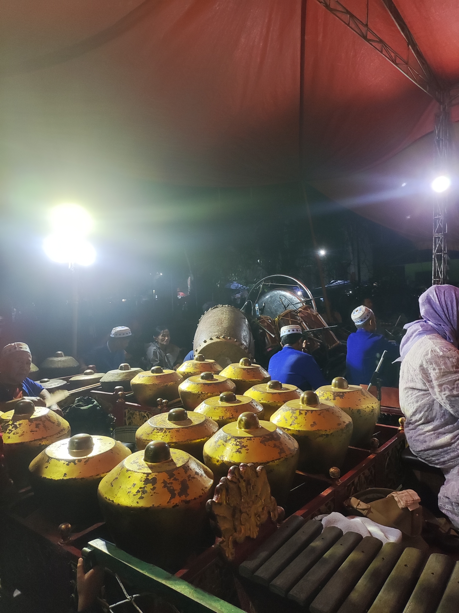 The image shows a gamelan performance taking place at night under a red canopy. In the foreground, rows of golden gong instruments with prominent knobs are visible. Several individuals, predominantly men wearing peci caps and modest attire, are seated behind the instruments, appearing to be musicians. One person on the left is actively playing a gong. In the background, a large drum and other percussion instruments are present, along with a few more individuals watching the performance. A woman wearing a light purple hijab is visible on the far right, observing the scene. The lighting is dim, with bright spotlights illuminating parts of the stage and the instruments. The overall atmosphere suggests a cultural event or performance. The location is identified as Panongan, Indonesia.