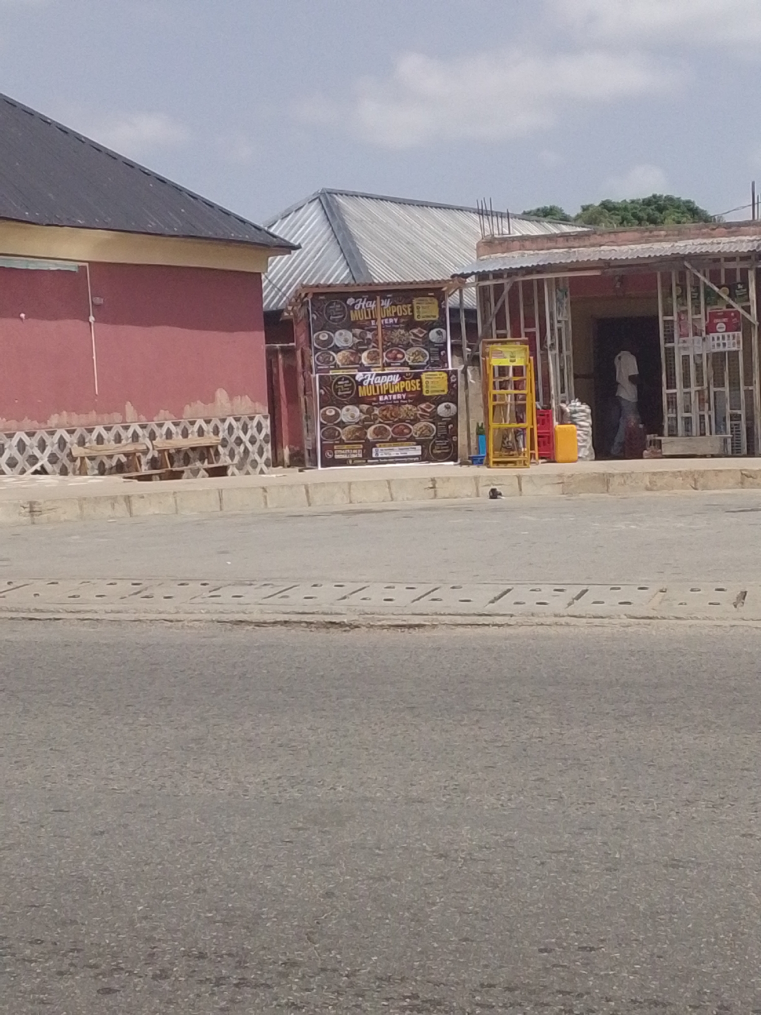 The image shows a street scene in Jalingo, Nigeria, on a bright, clear day. In the foreground is a paved road with drainage grates. Behind the road, there is a sidewalk with a patterned border.
On the sidewalk, there are several structures. To the left is the side of a building with a red-pink wall and a dark grey metal roof. Next to it, there is a simple wooden bench placed near a decorative tiled wall.
Further to the right, there is a small eatery or shop with an open front. A sign advertising "Happy Multipurpose Eatery" is prominently displayed, featuring images of various dishes. Beside the entrance, a person in a light-colored shirt and dark pants is standing, and there are some stacked items, including a yellow container and some bags. A yellow rack with shelves is also visible. The overall setting appears to be a commercial area with shops and buildings lining the street. The sky is blue with some clouds, suggesting it is daytime.