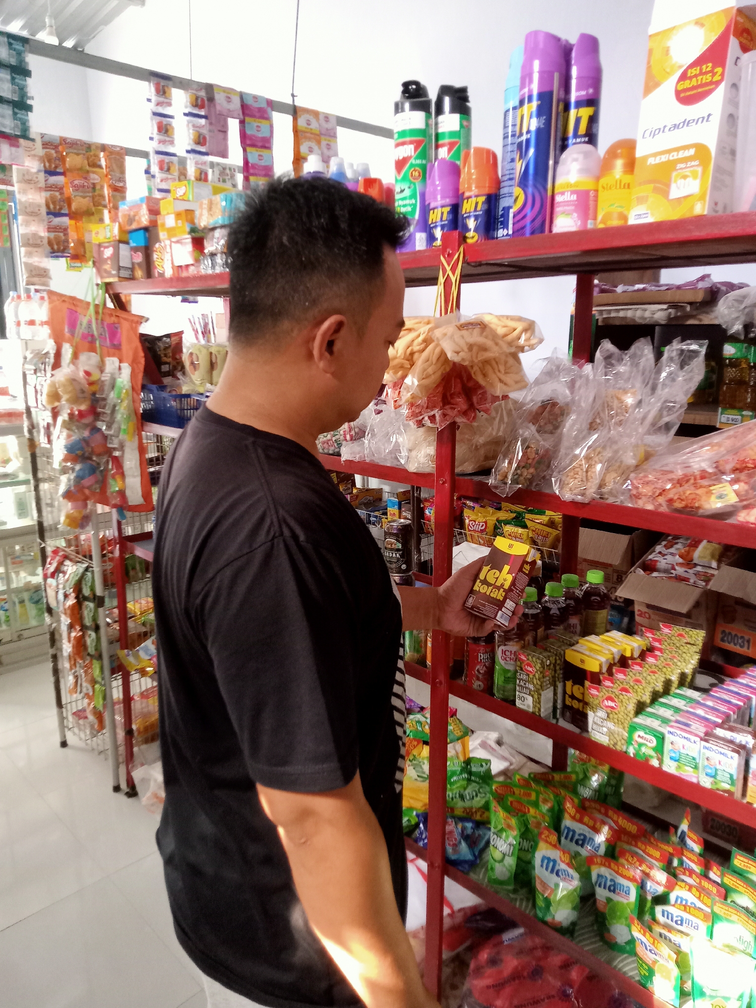 In Simo, Indonesia, a man in a dark t-shirt is standing in a convenience store aisle, looking at a shelf of goods. He is holding a yellow and brown box in his right hand, appearing to examine it. The shelves are stocked with various packaged snacks, drinks, and household items. The store appears to be well-lit, with fluorescent lighting overhead. Products visible include bags of crackers, bottles of beverages, and boxes of instant noodles. A metal display rack is present on the left side of the image.