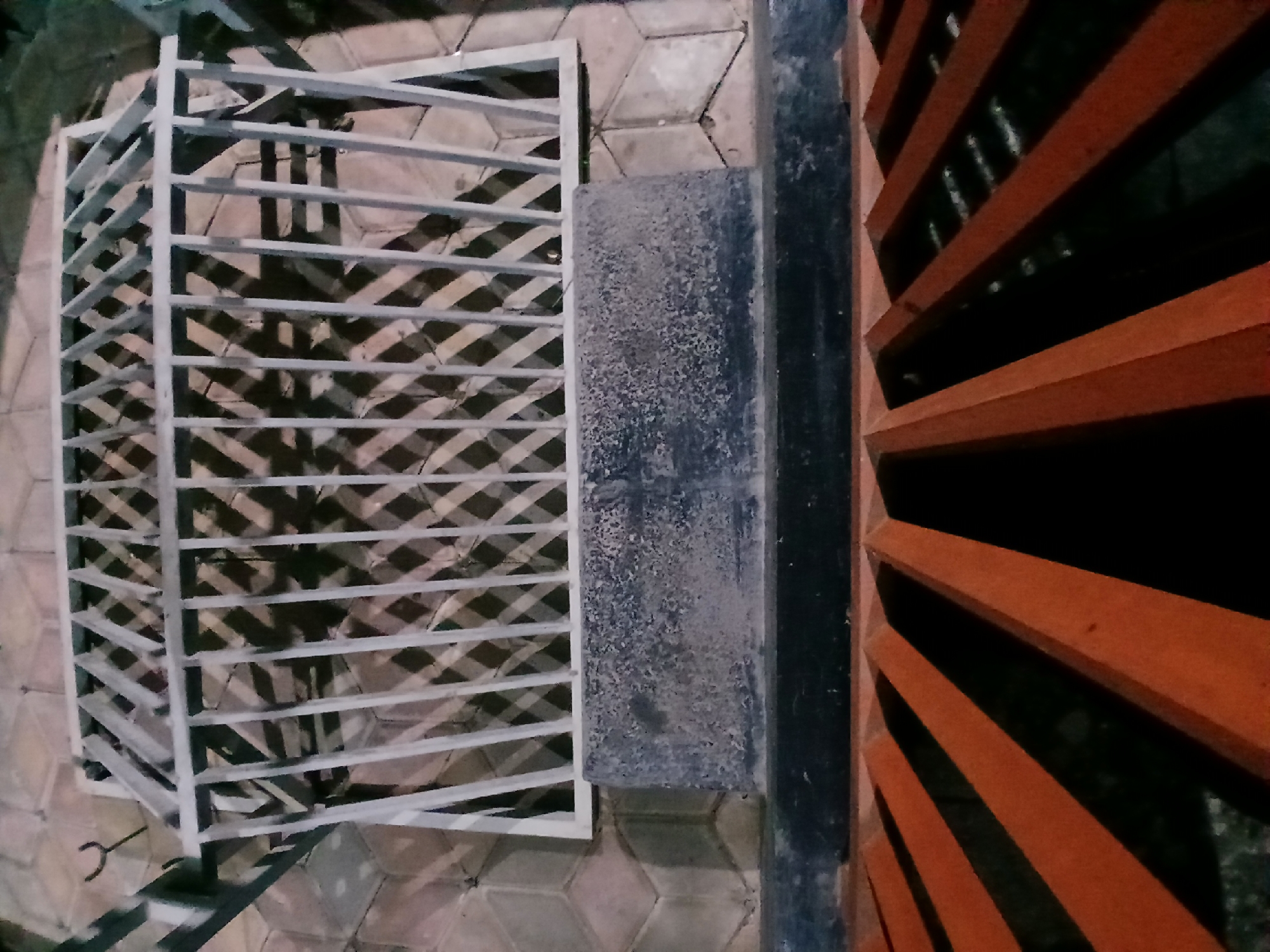 The image shows a close-up, overhead view of a metal grate. The grate is made of white metal bars arranged vertically and diagonally, creating a diamond pattern. It appears to be a decorative element, possibly part of a fence or a window covering. The grate is casting shadows on the ground, which is paved with hexagonal tiles. Below the white grate, there is a rough, dark grey concrete structure. At the bottom of the image, a section of an orange metal railing is visible, with diagonal bars creating a V-shaped pattern. The overall scene suggests an outdoor urban environment, potentially in Sekarputih, Indonesia, given the context provided. There are no people or animals visible in the image, and no actions or interactions are depicted. The lighting suggests it might be evening or nighttime, as shadows are prominent and the overall illumination is somewhat low.