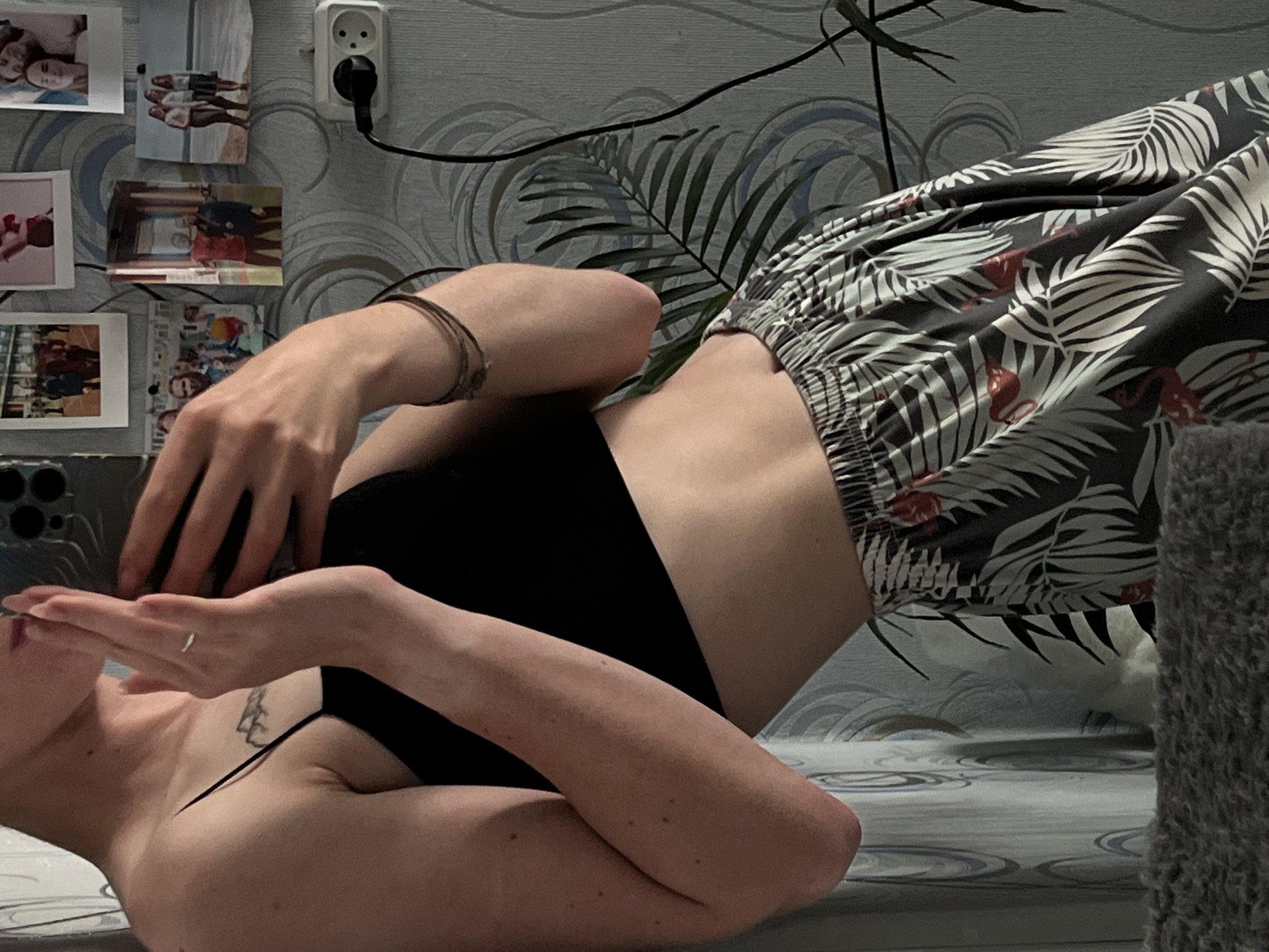 The image depicts a person indoors, likely in a domestic setting in Chaikovsky, Russia. The person is leaning against a wall adorned with floral wallpaper and several framed photographs. They are wearing a black crop top and patterned pants. The subject is holding a smartphone, suggesting they are taking a selfie or recording a video. Behind them, an electrical outlet is visible, with a cord plugged into it. A houseplant with large leaves is also present in the scene. The overall impression is of a casual, personal moment captured within a room.