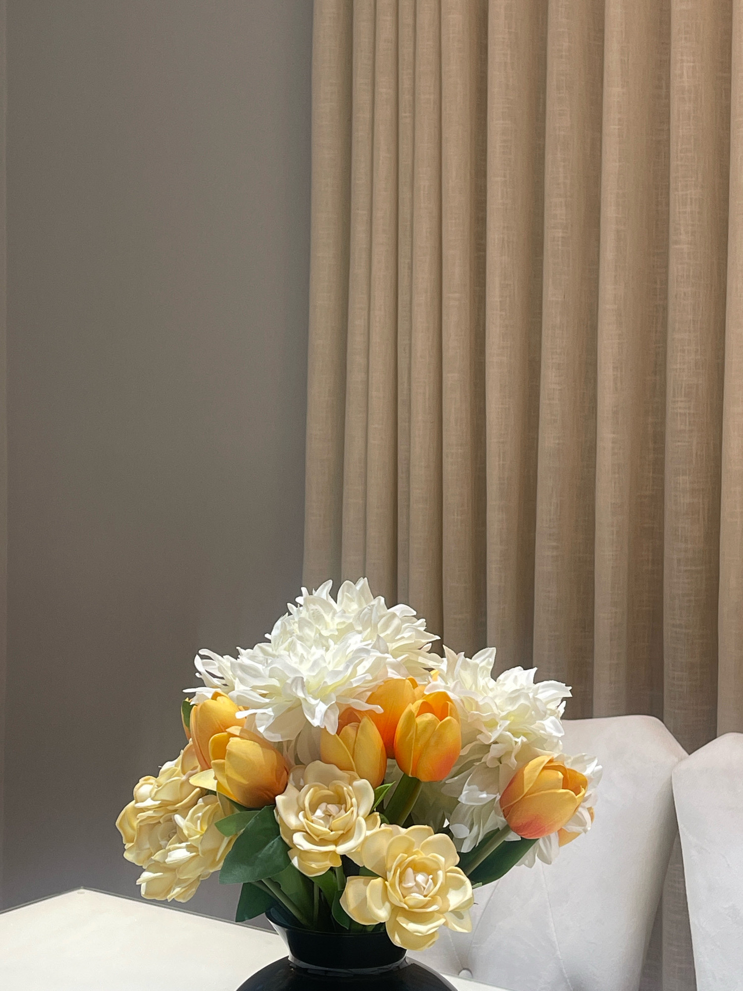 A bouquet of artificial flowers, predominantly in shades of yellow and white with some orange tulips, is arranged in a black vase on a white surface. The arrangement is positioned in the foreground, slightly to the left of center. Behind the flowers, cream-colored curtains with vertical pleats are visible. To the right of the curtains, two white upholstered chairs are partially visible. The background also includes a plain grey wall on the left side of the image. The scene appears to be set in an indoor residential or hospitality setting in Jabriya, Kuwait. There are no people present in the image.