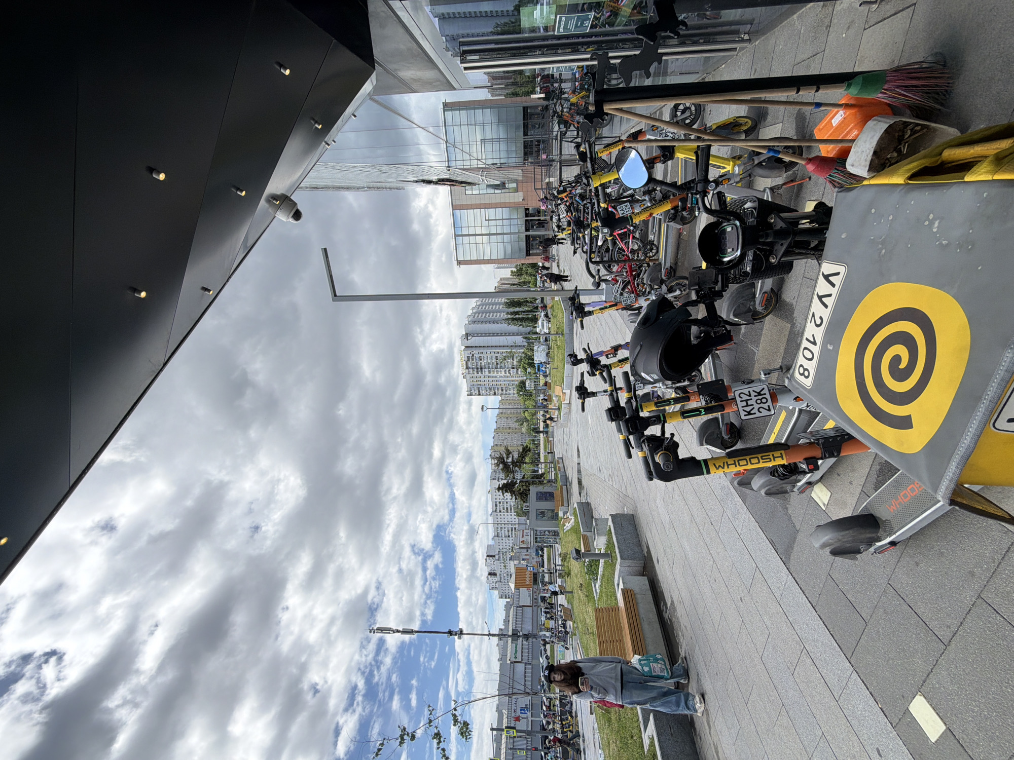The image captures an urban scene in an unspecified city. A gray pavement runs along a building, where numerous bicycles and scooters are parked. Many of them are marked with "WHOOSH." In the foreground, one scooter with a yellow spiral design on a gray panel is visible, with the license plate "KH2 28K." Brooms and cleaning equipment are haphazardly placed among the bikes. A woman, wearing jeans and a gray top, stands near a bench with a bag of groceries. In the background, residential buildings are visible under a cloudy sky. The architectural style of the buildings suggests a contemporary or post-Soviet era. The photo is taken from an elevated perspective, tilted to the side.