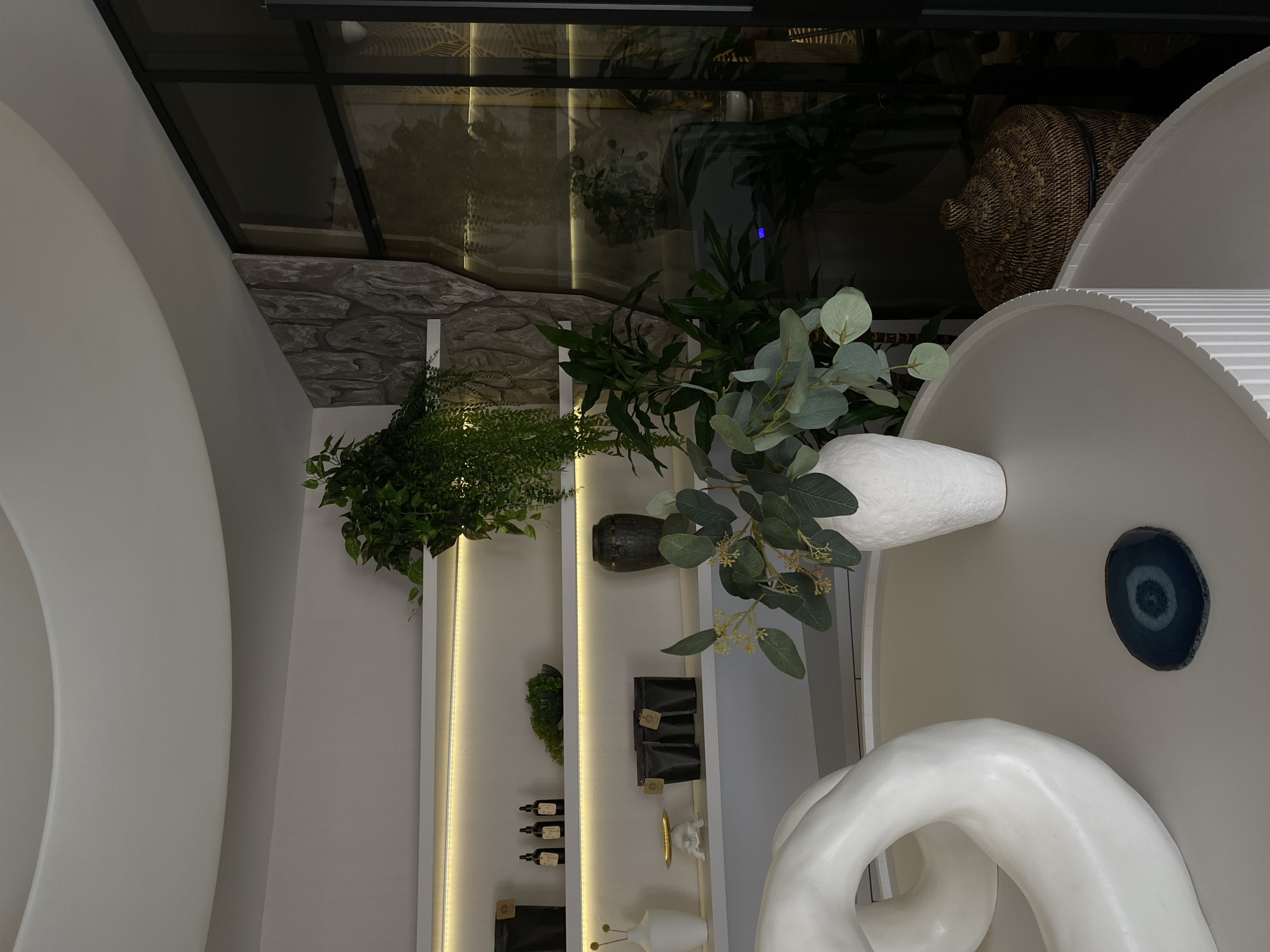 A still-life arrangement is featured in a well-lit room. Shelves are visible with a variety of decorative items. A green plant cascades down from the top shelf. A white vase holds greenery. There is a white, circular sculpture and a decorative piece on the wall. The ceiling has a glass and stone design. A basket is partially visible in the background. This scene is not necessarily located in Votkinsk, Russia.