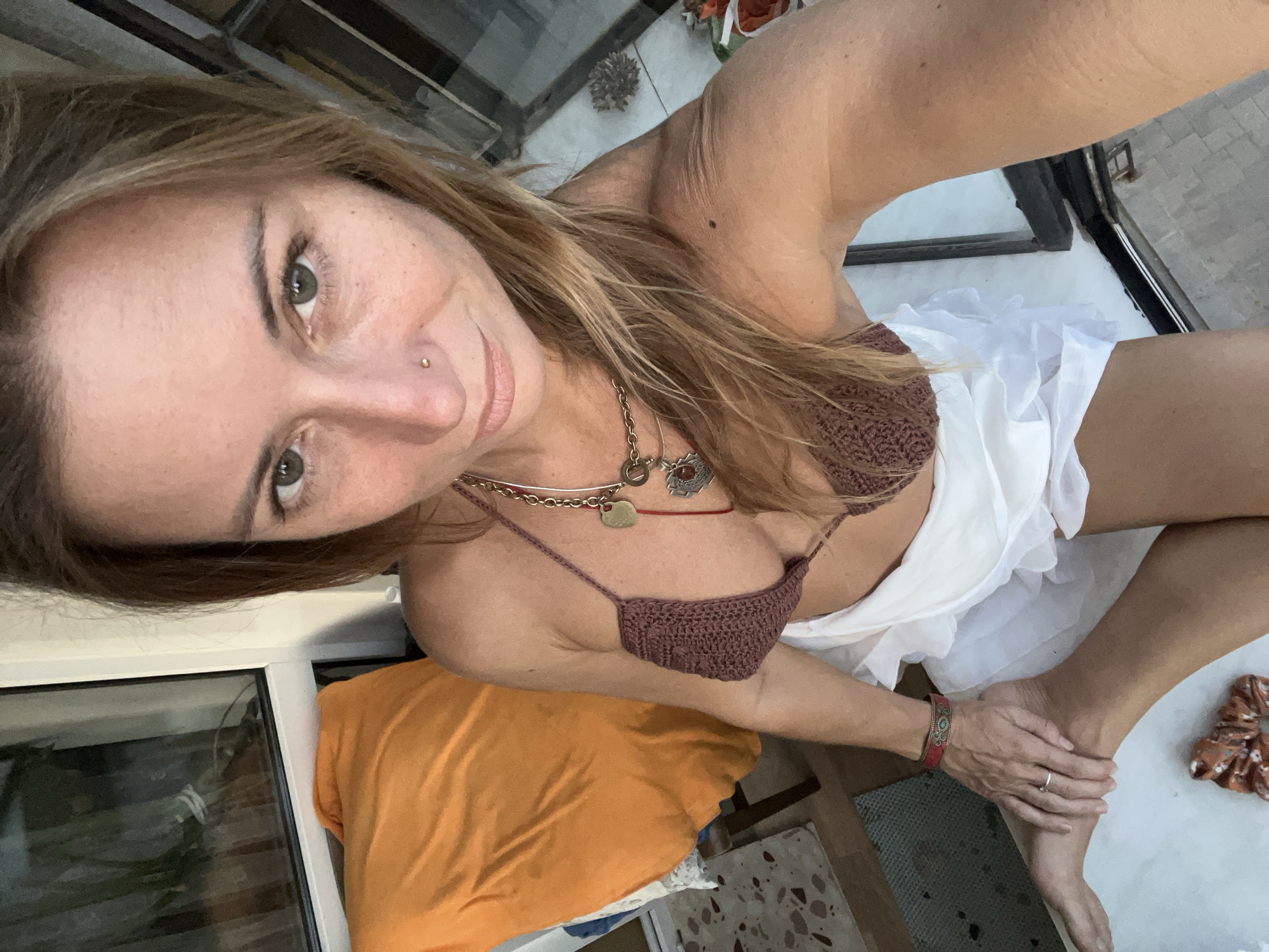 A woman with blonde hair and green eyes is sitting on a windowsill. She is wearing a brown crocheted bikini top and a white skirt. Her left hand is resting on her left knee, and her right arm is extended, likely holding the camera. Behind her, an orange pillow is visible on a chair next to a window. The scene appears to be an indoor or balcony setting, possibly in Samara, Russia, given the context provided. The lighting suggests it is daytime.