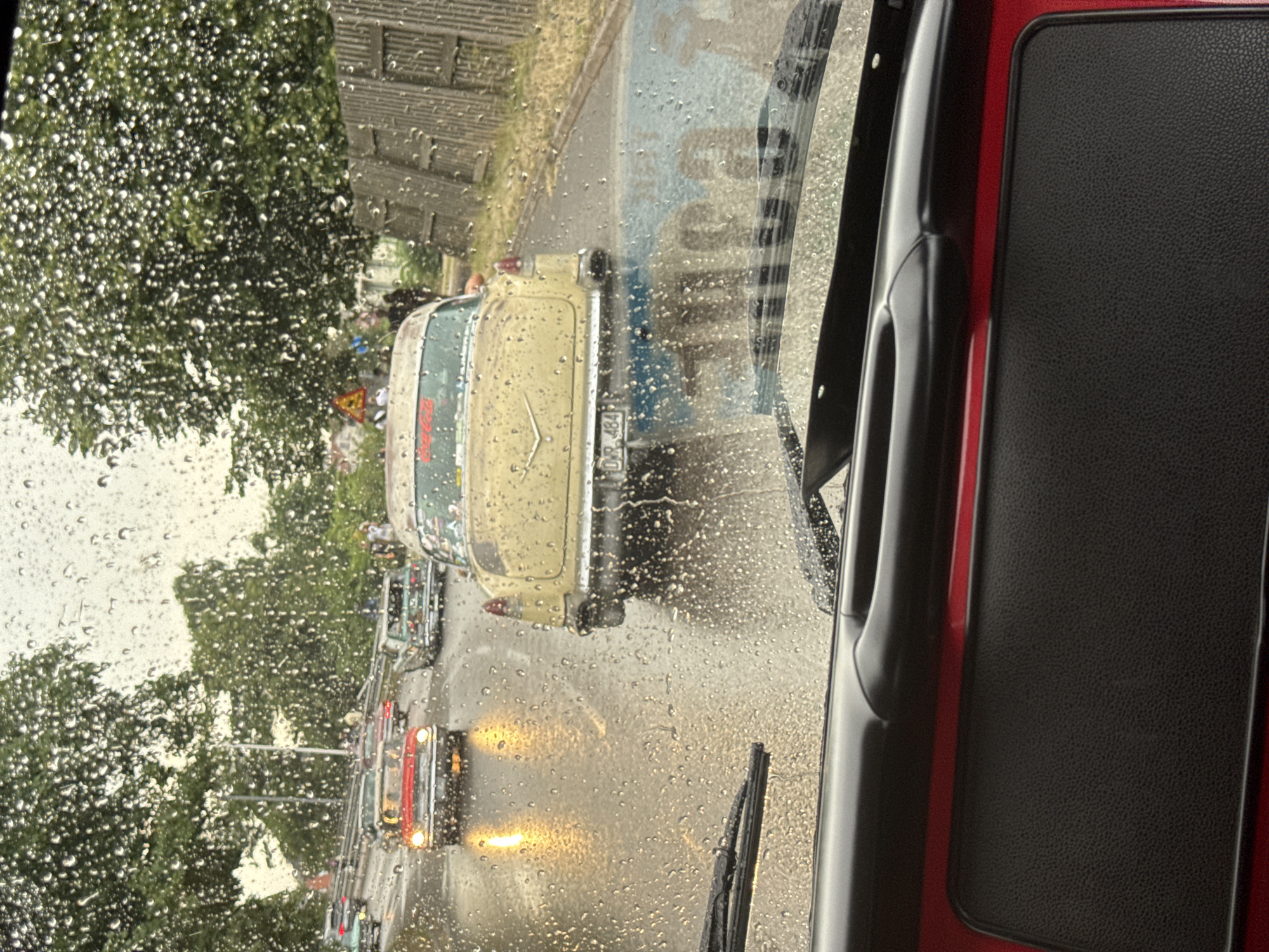 The image captures a view from inside a vehicle looking out through a rain-streaked windshield. It appears to be a procession or gathering of vintage cars on a wet street. The dominant subject is a yellow vintage car in the foreground, viewed from the rear, with the license plate "DRK 484" visible. To its left, a red vintage car is visible with its headlights on, reflecting on the wet asphalt. Further down the road, other vintage cars are present, and several people are standing on the roadside. The environment suggests an overcast day with rain, indicated by the water droplets on the windshield and the wet road surface. The presence of trees lining the street and a wooden fence on the right side of the road suggest an urban or suburban setting. Given the context of a vintage car event and the specific license plate, this scene is likely taking place in Falkenberg, Sweden, a location known for such gatherings.