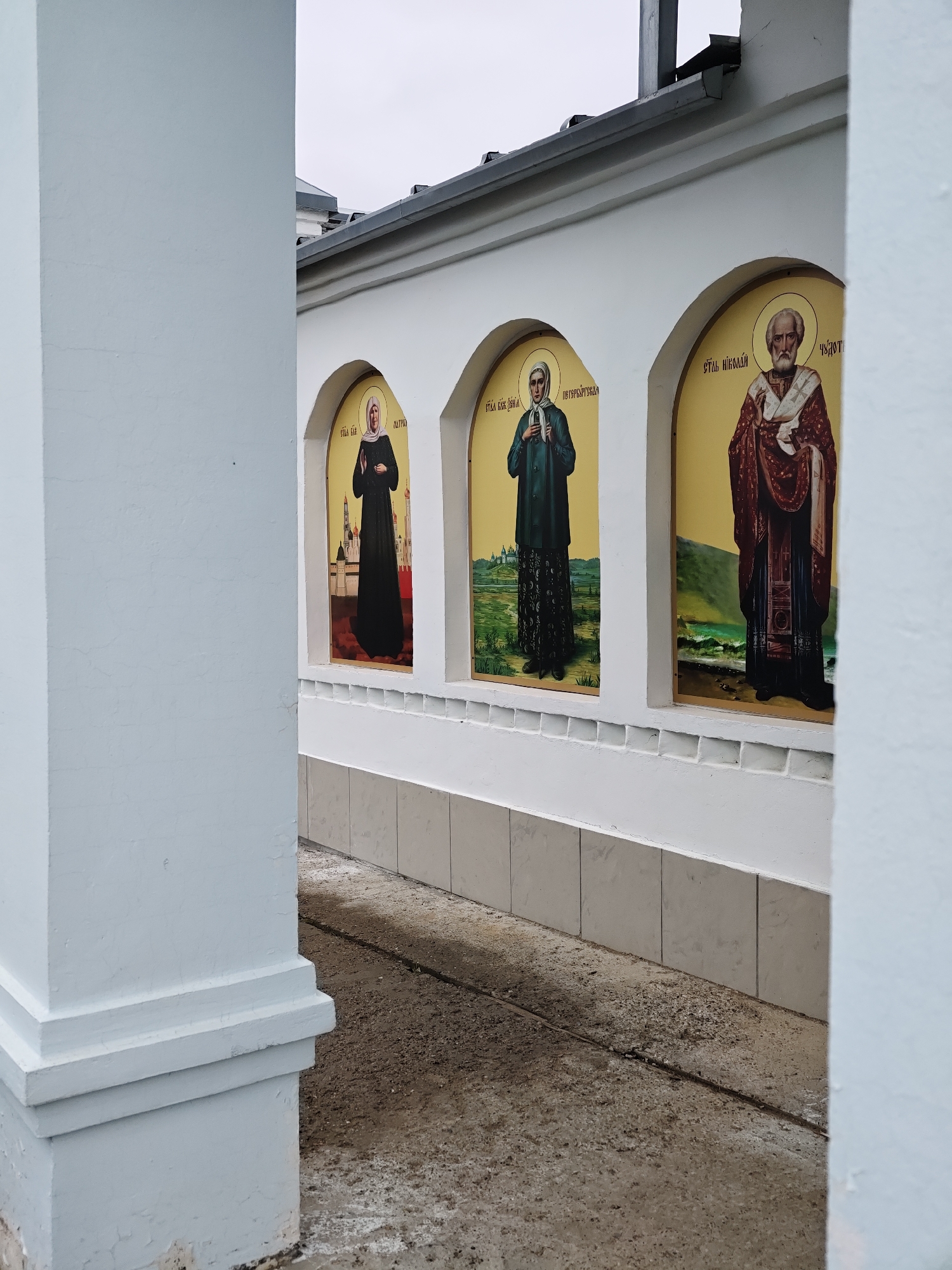 The photograph depicts a section of a white building exterior, possibly a monastery or religious structure, situated in Noviy, Russia. The perspective is framed by a white pillar on both the left and right sides. Embedded within the wall are three arched niches, each containing a religious icon featuring full-length figures. The first icon from the left displays a woman in a dark habit with a white headscarf, the second shows a woman in a dark green habit with a patterned skirt and a white headscarf, and the third presents a bearded man in red and white robes. Russian inscriptions are above each figure. The wall has a tiled base. The foreground shows a concrete walkway. The sky appears overcast.