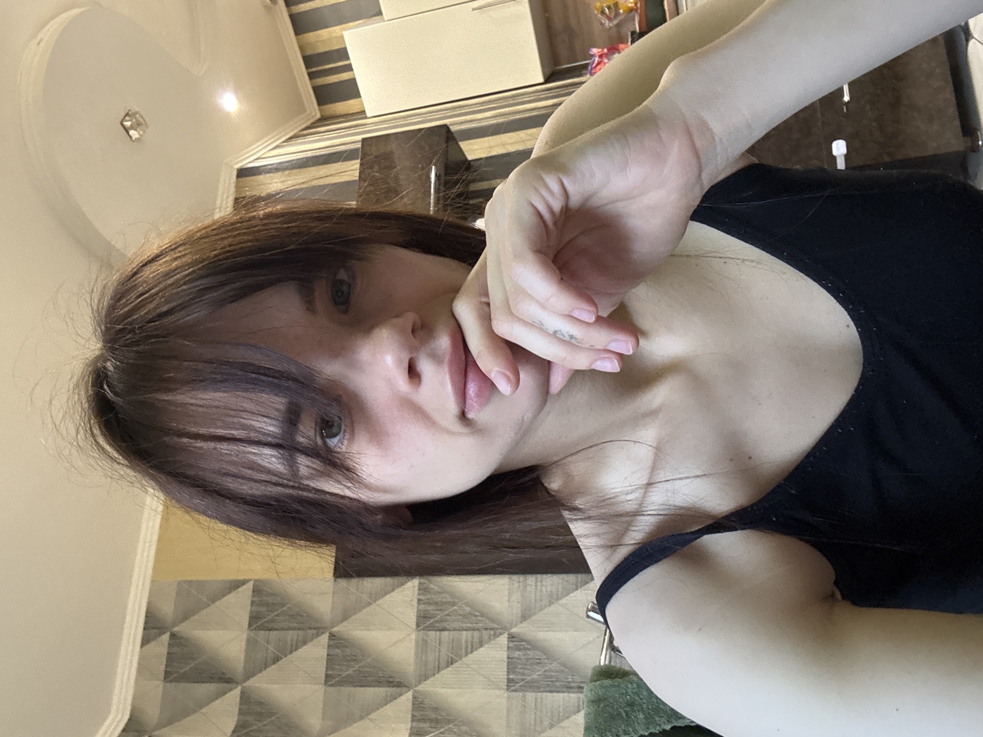 The image is a rotated, close-up selfie featuring a young female. She has long, dark brown hair with bangs that partially obscure her forehead. Her light-colored eyes, possibly green or hazel, look directly at the viewer. Her expression is neutral, with her lips slightly parted in a subtle pout. Her left hand is raised, with fingers gently resting near her chin and mouth. A small, indistinct tattoo or marking is visible on one of her fingers. She wears a plain black tank top or camisole, part of which is visible around her shoulder and upper chest.
The setting is indoors, likely a room in a home. The background includes a wall covered in a distinct geometric patterned wallpaper with shades of gray, brown, and cream, creating a textured, almost three-dimensional effect. Adjacent to this, a solid light-colored wall is visible. On the ceiling, a decorative white light fixture in the shape of a heart, featuring a small reflective element in its center, is present. A light-colored cabinet or shelf unit can be seen in the upper background, holding a few small, indistinct objects, including some colorful packaging. A dark, possibly reflective, surface is also visible in the background. A small portion of a green, textured object, potentially a decorative item or part of a bag, is visible at the bottom right.
The lighting suggests a well-lit environment, possibly daytime or bright indoor artificial light. The overall impression is that of a casual, posed portrait. While not depicted visually, the provided context places this scene in Rostov-on-Don, Russia.