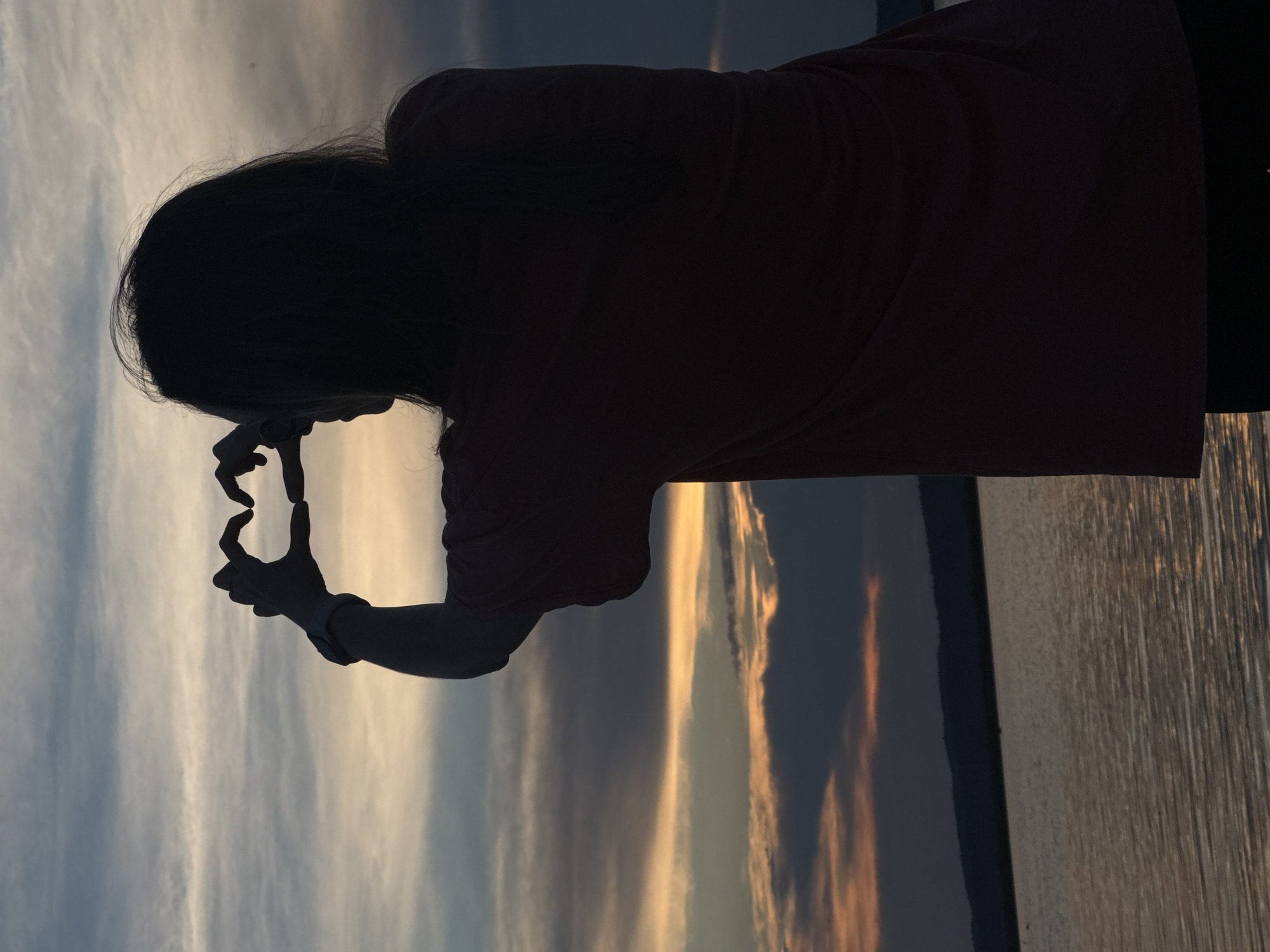 In this silhouette image, a person stands with their back to the camera, forming a heart shape with their hands in front of a dusky sky. The person appears to be on a waterfront, with the calm water of the Kama River reflecting the subdued colors of the sunset. The sky is filled with bands of pale yellow, orange, and gray clouds, suggesting the late evening. In the distance, a dark, undulating silhouette of hills or mountains can be seen against the horizon. The person is wearing a t-shirt and has long hair. This scene is captured in the city of Chaikovsky, Russia, likely at a scenic overlook or a park along the Kama River.