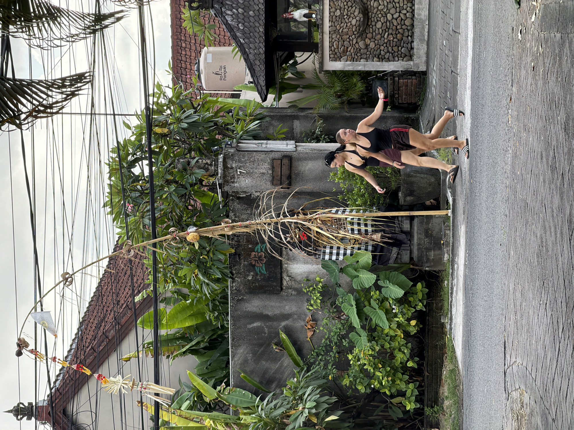 In Kuta, Indonesia, two young women walk on a paved street past a grey concrete wall and lush green vegetation. The woman on the left wears a black tank top and a maroon skirt, while the woman on the right wears a black tank top and brown shorts with red trim. They are walking from left to right, away from the camera.
To the left of the women is a tall, thin bamboo pole adorned with colorful decorations and a white flag. Behind the wall, a tiled roof and electrical wires are visible, suggesting a residential area. To the right of the women, a building with a stone facade and a dark tiled roof stands, with a statue of a rooster visible inside. The overall scene captures a street in Kuta, Indonesia, with elements of local culture and everyday life.
