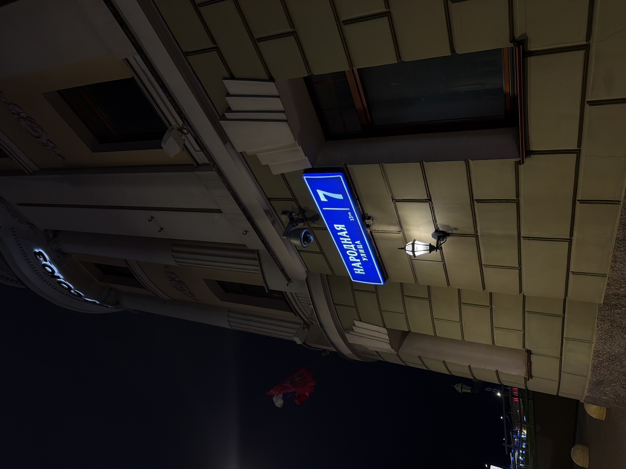 Here is a description of the image:
The image shows a street at night, likely in an urban area. The focus is on a building facade, which includes a blue street sign. The street sign has white lettering and the number "7" on it. The sign reads "НАРОДНАЯ УЛИЦА" in Russian, which translates to "People's Street". There is a street light fixture mounted near the sign. A surveillance camera is also attached to the building near the sign. Below the street sign there is a small red flag with white and blue accents, possibly a national flag. The building itself has a decorative architectural style with a brick-like pattern on the exterior walls. A window is visible in the background, reflecting the night sky. In the far background there is indistinct urban scenery.