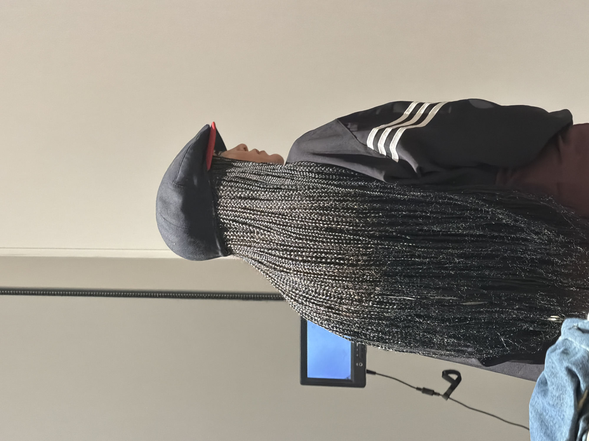 A person with long, black braids is lying on a surface. They are wearing a black Adidas jacket with white stripes on the sleeves, and a black cap with a red brim. A small electronic device is positioned beneath the person's braids. The background is a plain wall. There is no information present to suggest the city is Yerevan, Armenia.