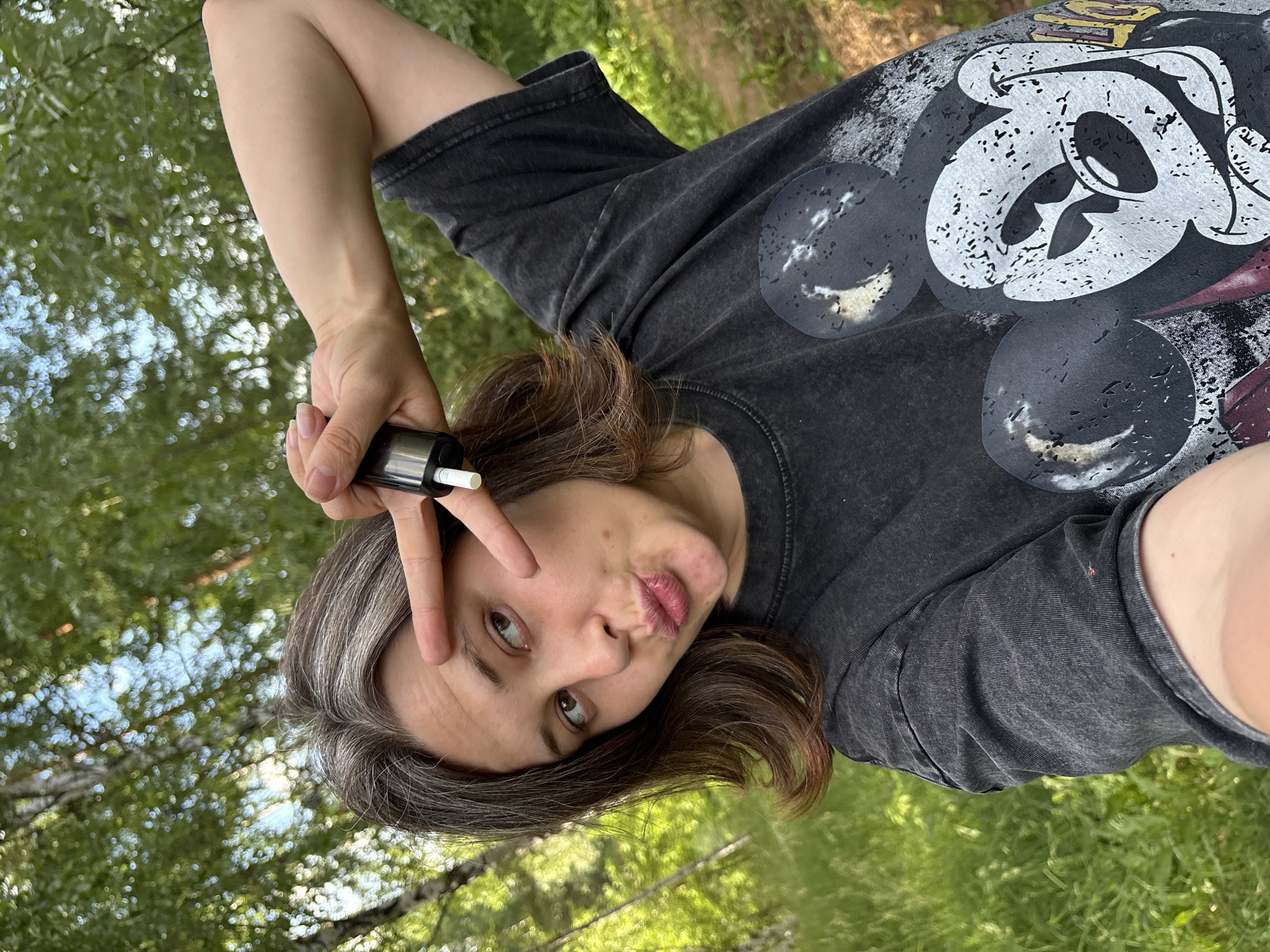 In this vertical, medium shot, a young person is positioned in a natural, outdoor setting with lush green foliage in the background. They are wearing a dark gray t-shirt with a faded graphic print of Mickey Mouse. The person is holding a black cylindrical object in their right hand, which appears to be a vape pen or similar device, with a white tip visible. Their left hand is raised, forming a peace sign gesture near their face.
The individual has medium-length, dark brown hair with streaks of gray. They are looking directly at the camera with a playful expression, puckering their lips. The lighting suggests it is daytime, with the sun filtering through the trees, creating a dappled effect. The overall atmosphere is casual and relaxed, possibly indicating a moment of leisure or a selfie-taking opportunity. The visual elements of the trees and greenery strongly suggest a park or wooded area. The provided location context of Chaikovsky, Russia, places this scene within that geographical area. No visible text is present in the image.