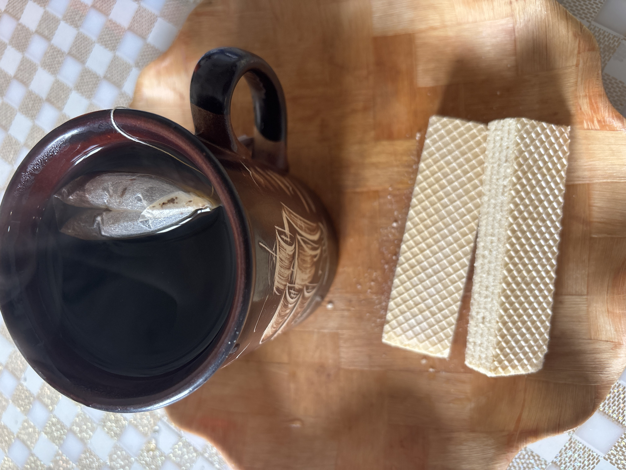 An overhead shot shows a mug of tea and two wafers on a wooden tray. The mug is brown with a pattern of a sailing ship. A tea bag is in the mug, which is filled with dark liquid. The wafers are light brown and have a grid pattern. The tray is on a checkered surface. The scene appears to be indoors, possibly in a home kitchen, with no indication of its location in Bila Tserkva, Ukraine.