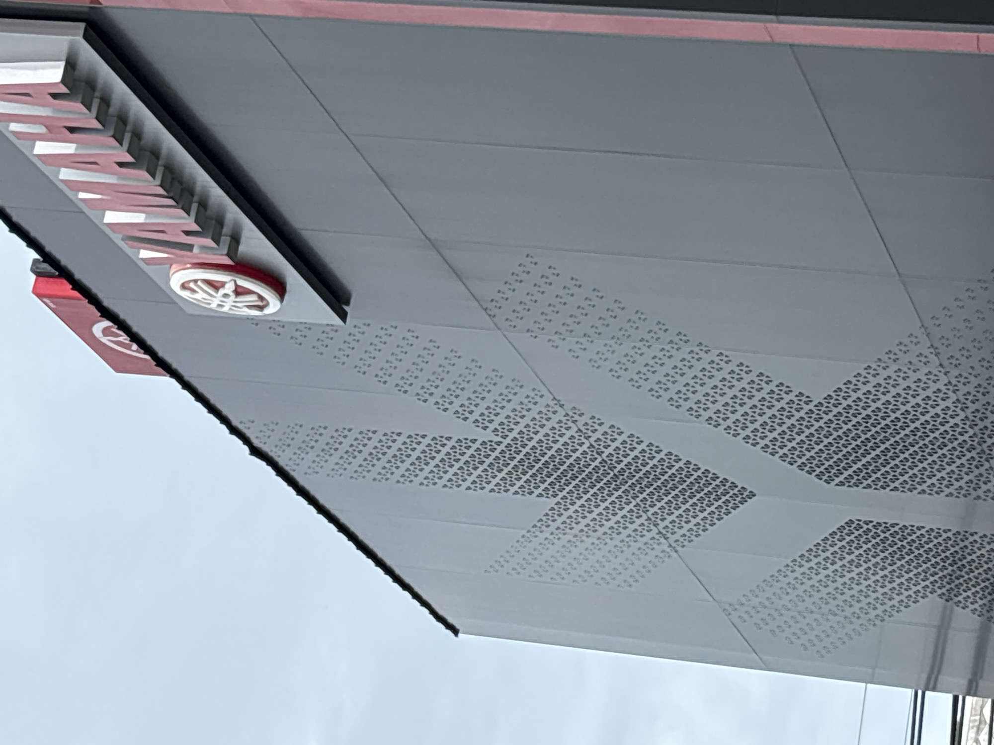 A partial, upward-facing view of a gray exterior wall with patterned cutouts and a "YAMAHA" sign in Baan Bo Phut, Thailand. The sign features white lettering outlined in red and a circular emblem. The wall is comprised of rectangular panels and has a red accent stripe at the top. The background shows a cloudy sky.