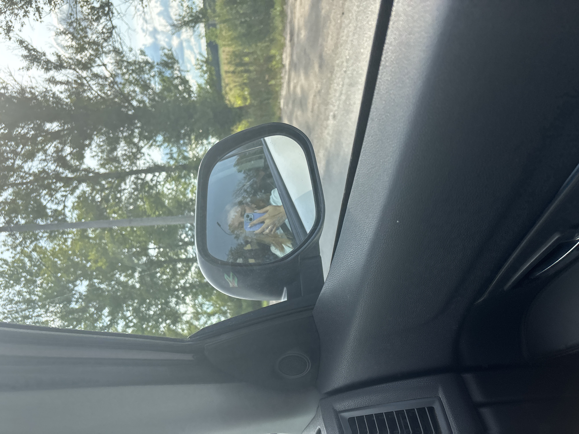 The image shows the interior of a car looking out the passenger side window. A side mirror reflects a young woman holding a smartphone and taking a selfie. The exterior scene visible through the window and in the mirror includes trees, a dirt road, and a cloudy sky. The interior of the car shows the door panel with a speaker and a door handle, as well as a vent. The image does not provide any specific information about the location in the city of Rusanovo, Russia.