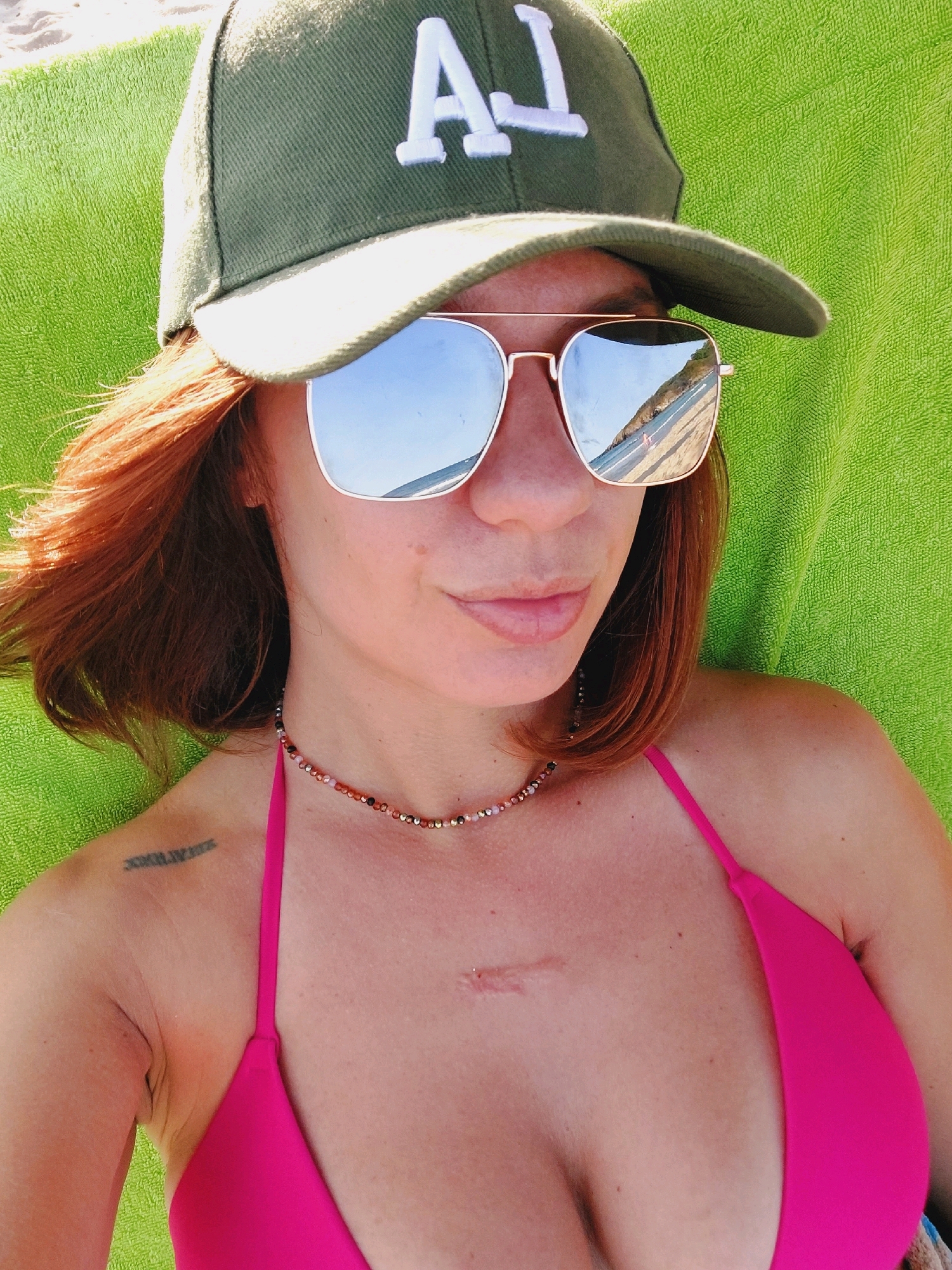 The image shows a woman lying on a green towel on a beach. She is wearing a green baseball cap with "LA" in white lettering, mirrored aviator sunglasses, a pink bikini top, and a beaded necklace. She has red hair and a tattoo on her left shoulder blade which appears to be Roman numerals. A scar is visible on her upper chest. The background is a beach with the sea and a hillside visible in the reflection of her sunglasses. The location is Sozopol, Bulgaria.