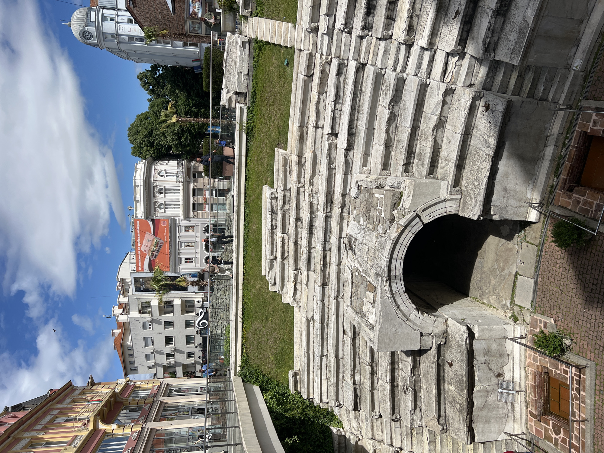 The image depicts the Ancient Theatre of Philippopolis in Plovdiv, Bulgaria. The Roman theatre's seating tiers are prominent, constructed of white stone. A circular entrance/exit is visible in the foreground. Behind the theatre, modern buildings rise, including a white building with a clock tower and another building with a large advertisement. The scene is situated in a city environment, as indicated by the surrounding buildings. People can be seen in the background behind a glass barrier. Grass and greenery are interspersed within and around the ancient structure. The sky is blue with scattered clouds.