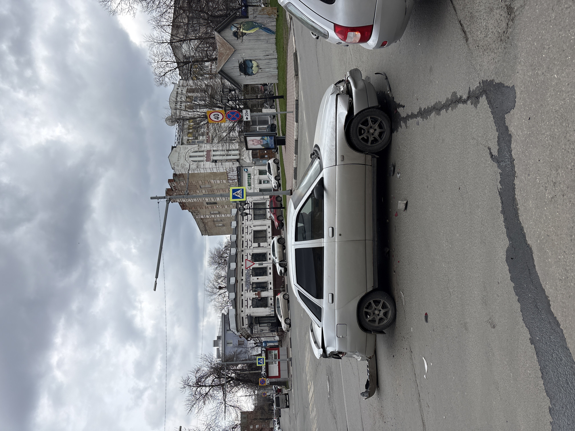 The image captures an urban street scene in Ulyanovsk, Russia, under an overcast sky, depicting the aftermath of a car accident. A light grey sedan is positioned on the right side of the road, exhibiting significant damage to its front-left fender, hood, and bumper area. Dark fluid, likely oil or coolant, has leaked from the damaged vehicle, forming a distinct trail on the asphalt that extends towards the bottom of the frame. The rear section of a second silver car is partially visible in the upper right, parked or stopped near the damaged vehicle.
On the left side of the street, several buildings are visible, including one with a striking mural of a parrot and another bird painted on its wooden exterior. Bare-branched trees line the sidewalks, suggesting the season is late autumn, winter, or early spring.
Various street signs are present, including a circular speed limit sign displaying "40," a "No stopping/parking" sign, and a blue square "Pedestrian crossing" sign. An advertisement stand features a person's face and the text "20М" (likely indicating 20 meters). Several Cyrillic signs are visible on the buildings, most notably a blue-crossed sign reading "АПТЕКА" (APTEKA), meaning "Pharmacy." The sky is uniformly cloudy, indicating an overcast daytime. The overall scene conveys a moment following an automotive incident in a city environment.