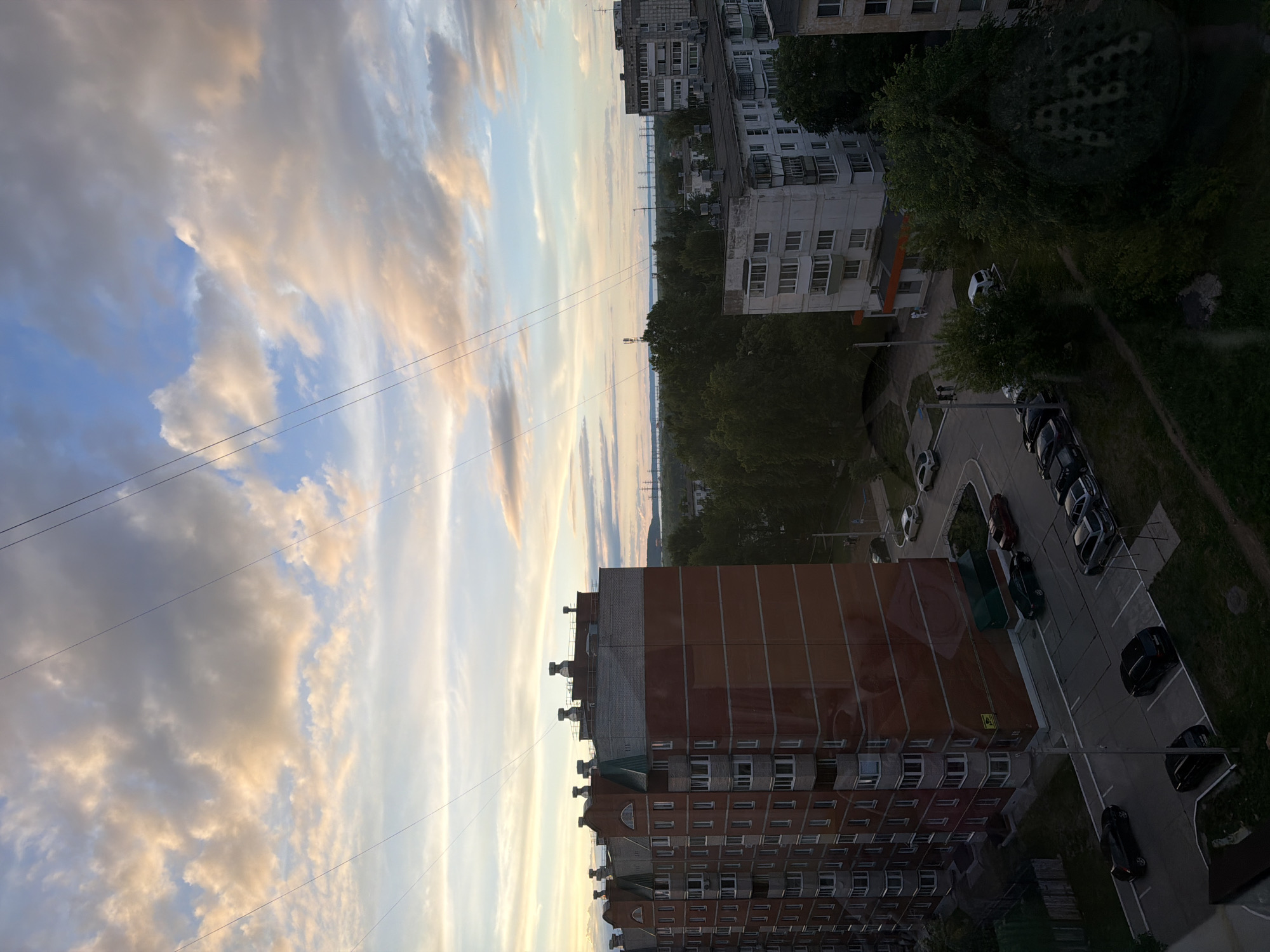 The image shows a high-angle view in what appears to be Chaikovsky, Russia, based on the architectural style of the buildings. The focus is on the sky, featuring a sunset or sunrise with clouds illuminated by the light. Lower in the frame, there are several multi-story residential buildings, indicative of an urban area. A parking lot with parked cars is also visible near one of the buildings. Trees are present along the edges of the built environment, suggesting a blend of urban and natural elements. Utility lines are visible stretching across the sky in the top left of the shot.