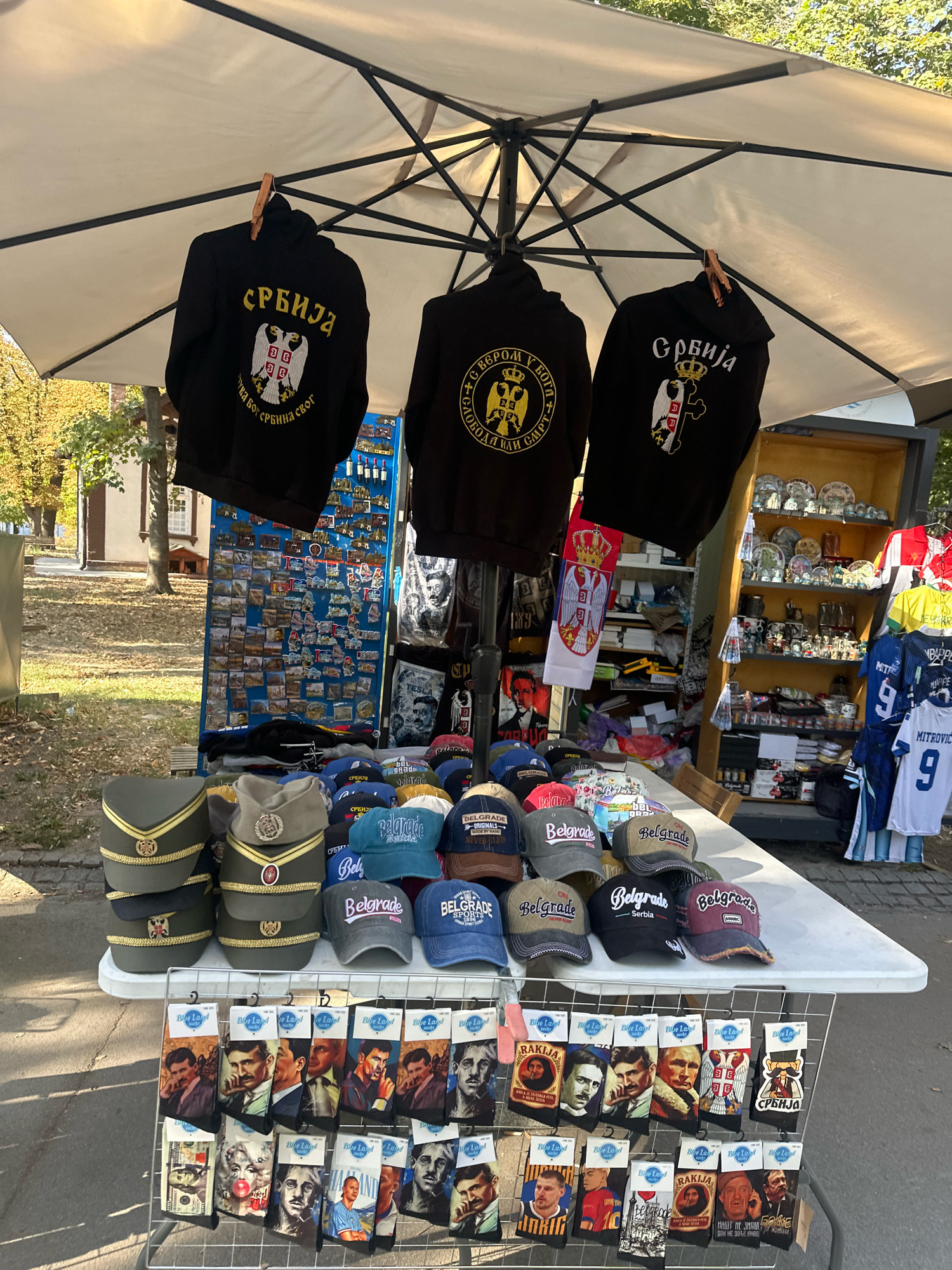 The image shows an outdoor souvenir stall under a large white umbrella. The stall displays a variety of merchandise, including black hoodies with Serbian flags and symbols, military-style caps in green and khaki, and a collection of baseball caps with "Belgrade" and Serbian flag motifs. In the foreground, there is a rack displaying socks featuring portraits of famous figures, currency, and Serbian motifs. Behind the caps and socks, a display board is covered in small souvenirs like magnets and postcards. The stall is set up on a paved area, with trees and buildings visible in the background, suggesting an outdoor market or tourist area in the city of Belgrade, Serbia. The overall scene depicts a commercial display of Serbian national and cultural memorabilia.