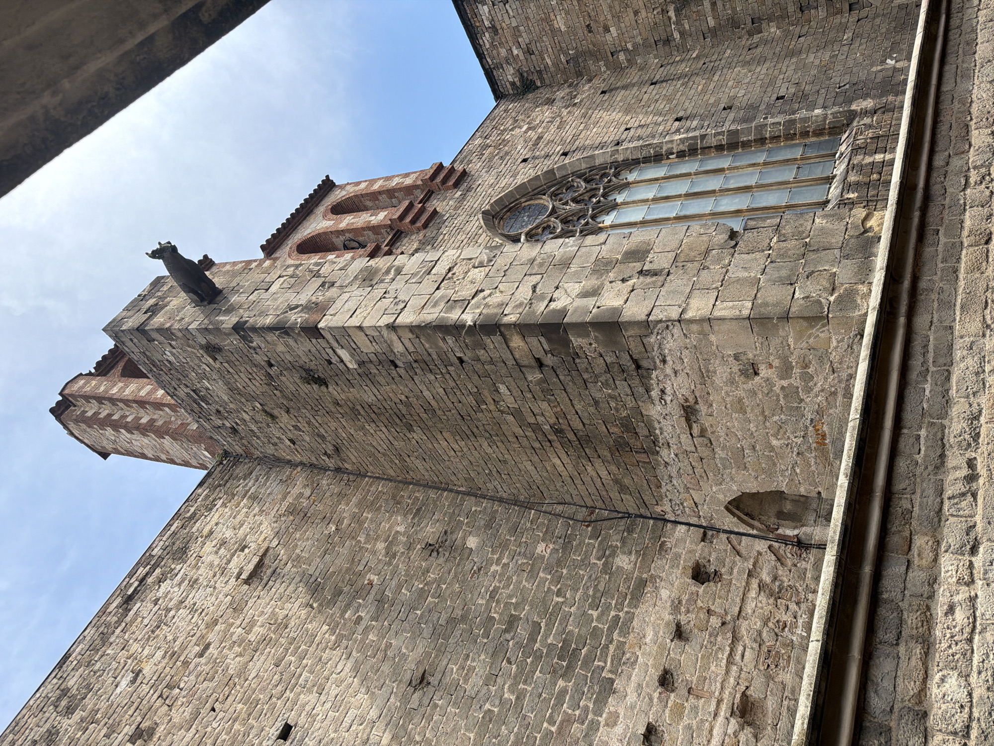 The image shows a low-angle view of a stone building, likely a church, with a sky that is partially obscured by a gray, angular structure in the upper right corner. The building is constructed from large, rough-hewn stones, giving it an aged and weathered appearance. A prominent feature is a gargoyle mounted on the side of a tower. To the right of the gargoyle, a tall, arched stained-glass window is visible. The window has a Gothic design with intricate tracery. The wall is adorned with a series of arched niches. A dark cable runs vertically along the stone facade. The location appears to be within the Gothic Quarter of Barcelona, Spain, a historic district known for its medieval architecture. The image is devoid of people or any visible activity.