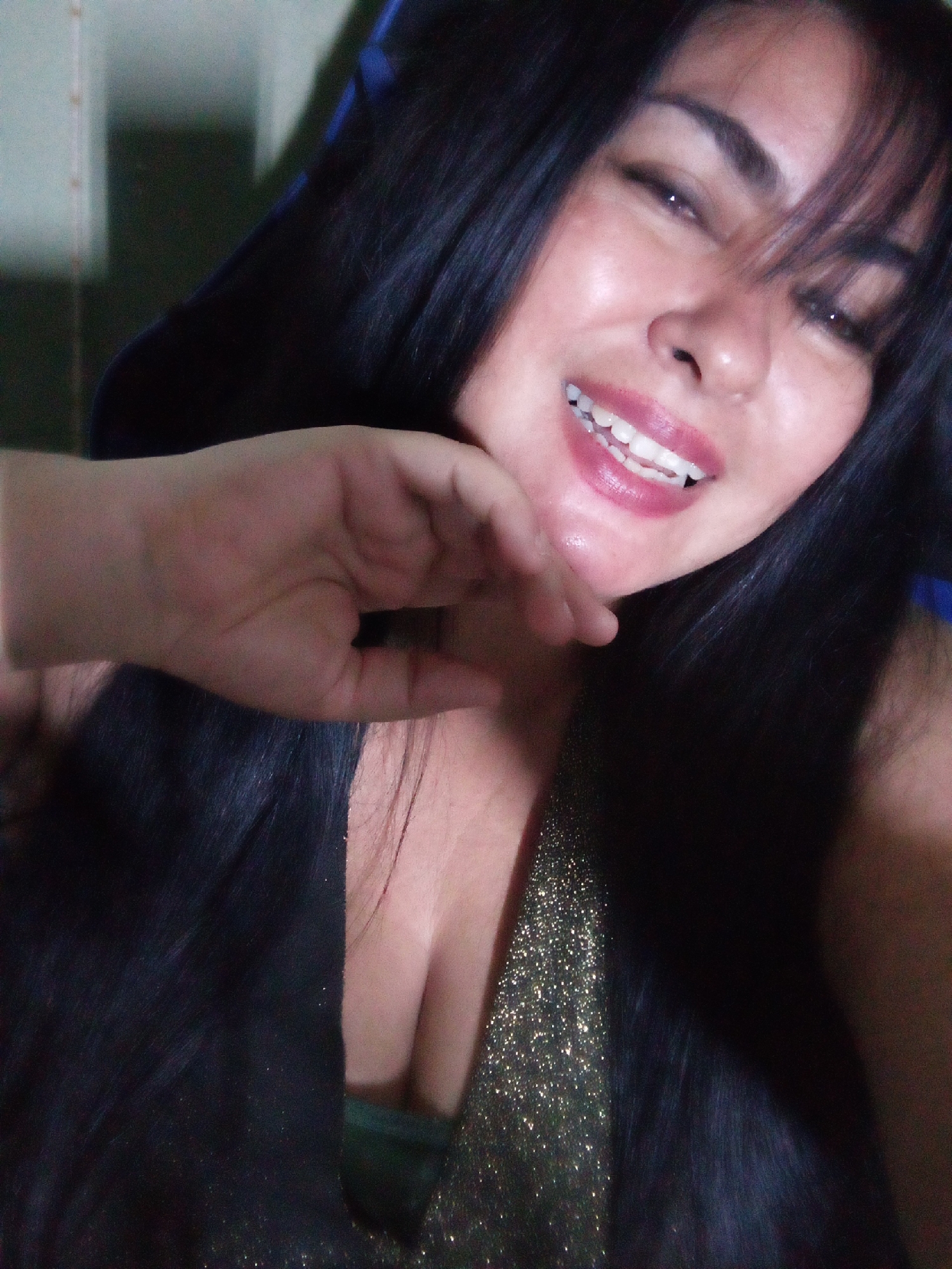 A woman is posing for a selfie. She has dark hair, dark eyebrows, and dark eyes. She is wearing a top that reveals part of her cleavage. The top appears to be sparkly on one side and dark on the other. Her hand is near her face and her mouth is open, showing her teeth. There is no information about the location.
