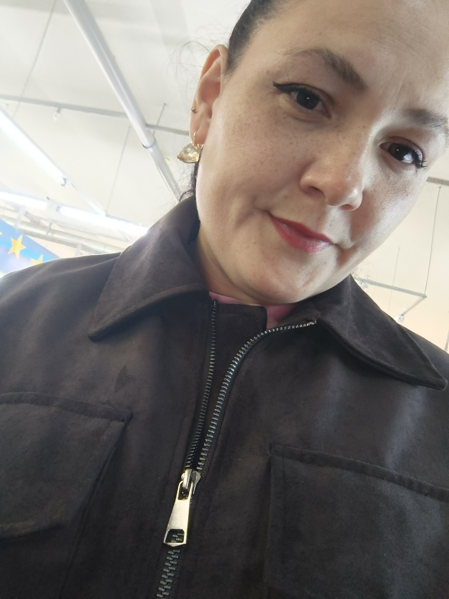 The image is a close-up, upward-angled selfie of a woman with dark hair, wearing a dark brown jacket with a zipper. She has dark eyeliner, lipstick, and a gold earring with a teardrop-shaped charm. The background is a white ceiling with fluorescent lighting and some pipes. A small portion of a blue banner with yellow stars is visible in the upper left corner. There is no information in the image to indicate the location in the city of Chaikovsky, Russia.