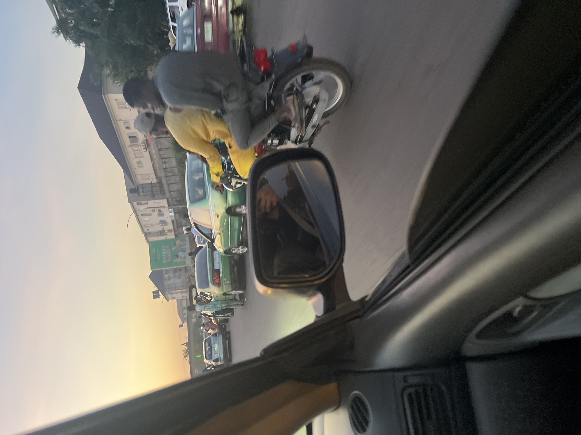 This photograph depicts a vibrant urban street scene captured from inside a vehicle during what appears to be the golden hour, as indicated by the warm, soft light illuminating the sky on the left.
In the foreground, the dark dashboard and a side mirror of the vehicle from which the picture was taken are visible. The side mirror reflects a blurred portion of the street and what seems to be a hand, likely the photographer's, holding a device.
The central subjects are two individuals on a motorcycle positioned next to the car. The driver, wearing a dark grey top, is seated on the motorcycle, while the passenger behind him is conspicuously dressed in a bright yellow top and trousers, looking towards the right. The motorcycle itself has silver-colored engine parts and wheels.
Beyond the motorcycle, a line of cars extends into the distance, suggesting traffic or a queue of vehicles. A green sedan is immediately visible next to the motorcycle, followed by other cars of various colors, including red/maroon and lighter hues. The road appears to be paved with asphalt.
In the background, multiple multi-story buildings line the street, featuring visible windows and diverse architectural styles. Some trees and foliage are also present above the buildings on the left side of the frame. A large green billboard or sign is partially visible in the distance, but any text on it is illegible due to resolution and distance.
The overall scene conveys a typical city street environment, bustling with vehicles and people, likely during a commute in an urban setting like Abuja, Nigeria. No discernible text is visible anywhere in the image. The weather appears clear, with no clouds or precipitation.