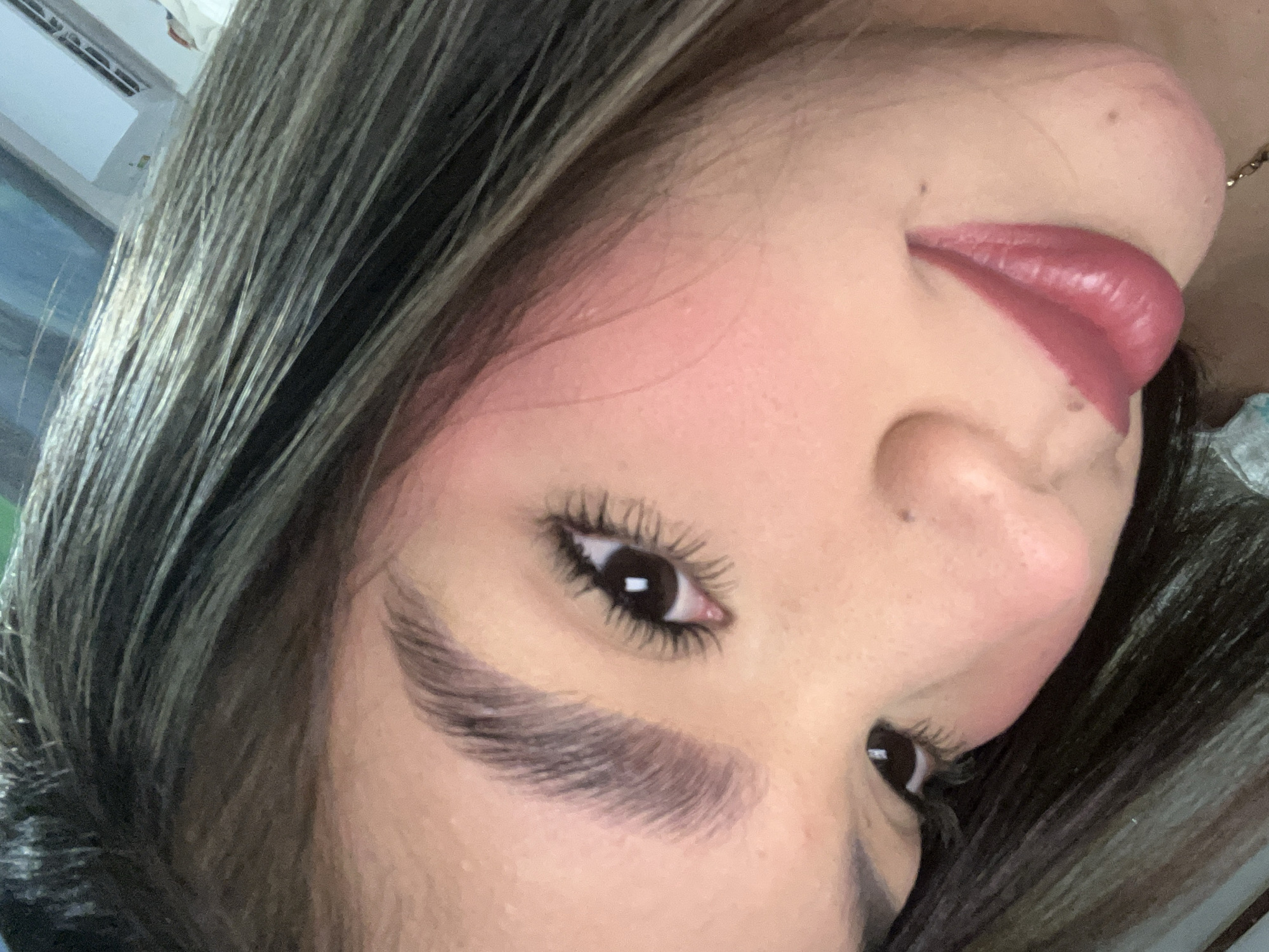 A close-up, eye-level portrait depicts a young adult female from the chest up, positioned with her head tilted to the right and looking slightly downwards. Her skin tone is medium with visible application of pink blush on the cheeks. Her dark brown hair, featuring lighter streaks, falls around her face and over her shoulders. Her eyebrows are thick, well-groomed, and brushed upwards. Her dark brown eyes are open, with prominent, volumized eyelashes, indicative of makeup application. A small dark mole is present on the right side of her nose. Her full lips are painted with a glossy, deep berry-red lipstick and are slightly parted in a gentle smile. In the upper right background, a white air conditioning unit is discernible, suggesting an indoor setting. No specific visual cues within the image identify the location as Juan Griego, Venezuela. A thin, gold-colored chain is partially visible near the bottom right edge of the frame.