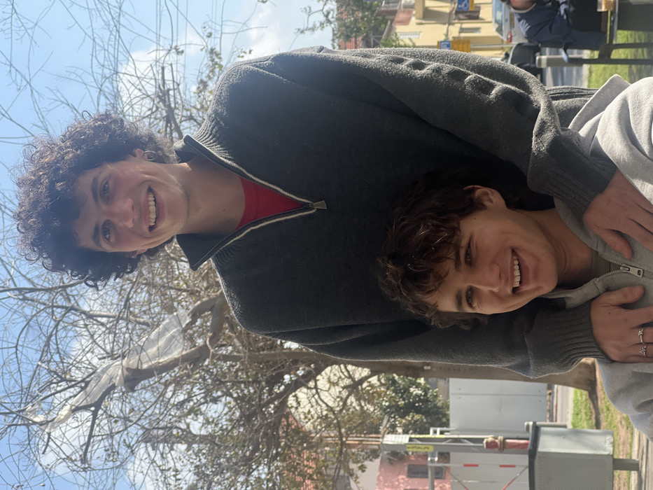 A daylight photograph captures two young adult males posing in an urban outdoor setting in Tel-Aviv, Israel. The taller male, with dark, curly hair and a subtle earring in his left ear, stands centrally in the background. He wears a dark grey quarter-zip sweater over a red shirt and smiles broadly, looking towards the camera. His hands rest on the shoulders of the shorter male, who is positioned in the foreground. This shorter male, with brown, wavy hair, wears a light grey zip-up hoodie over a green shirt. He also smiles broadly, looking slightly upwards and to the left. Two rings are visible on his left hand. His hands rest on top of the taller male's hands.
The background features bare deciduous trees under a clear blue sky with sparse clouds. A piece of white plastic sheeting is entangled in the branches of the prominent central tree. To the left, a light pink building is partially obscured by scaffolding, with a red fire hydrant and a grey utility box visible at ground level. To the right, another light-colored building, yellow street signs, and a distant male figure seated at an outdoor table with a beverage are present, consistent with a street scene in Tel-Aviv.