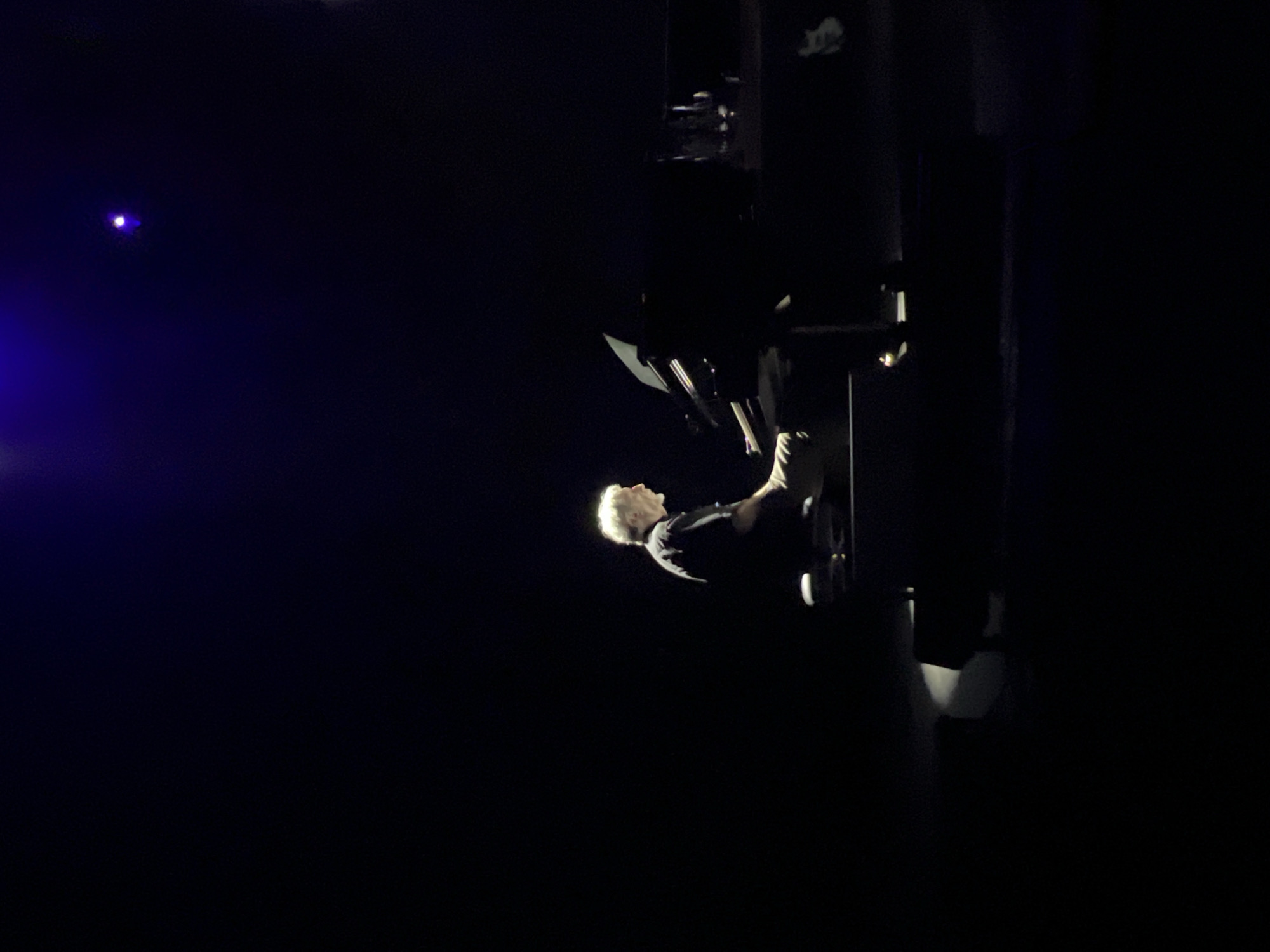 A man with short, white hair is seated at a black grand piano on a stage in Saint Petersburg, Russia. The stage is dark except for a single light illuminating the man. A blue spotlight is visible in the upper left corner. The man is wearing a black shirt.