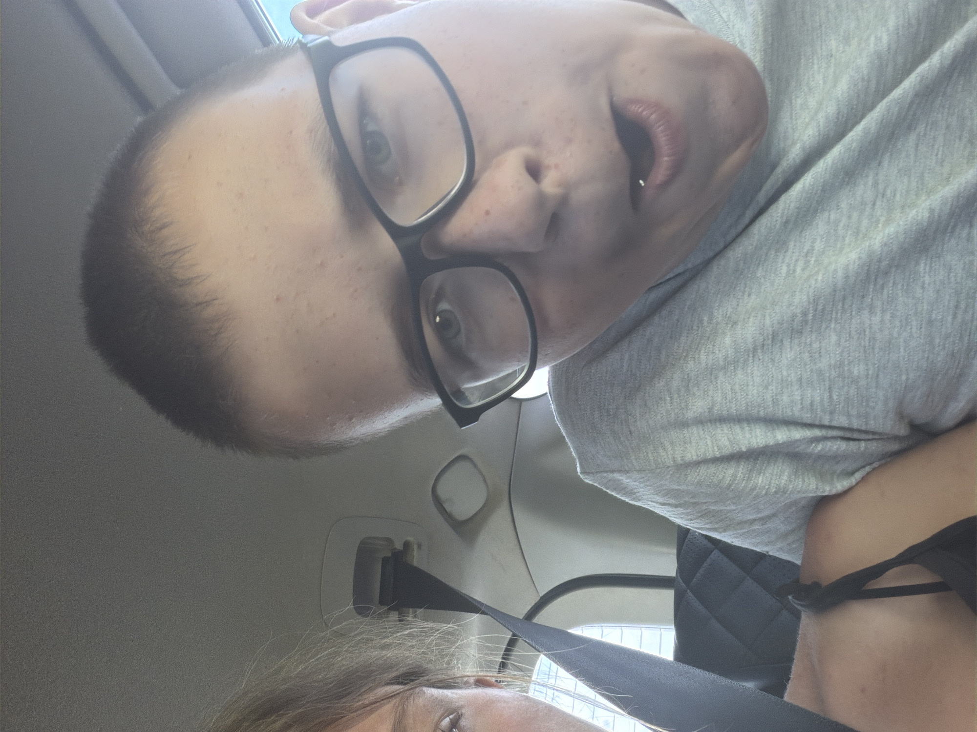 This is a selfie taken in a car. A person with short hair and glasses is in the foreground, making a surprised expression with their mouth open. Next to them, a person with blonde hair is partially visible on the left side of the frame. The interior of the car, including a seatbelt and a black padded seat, is visible. The image is lit by natural light, suggesting it was taken during the day. No specific details about the city of Ostrovka, Russia are provided in the image itself.