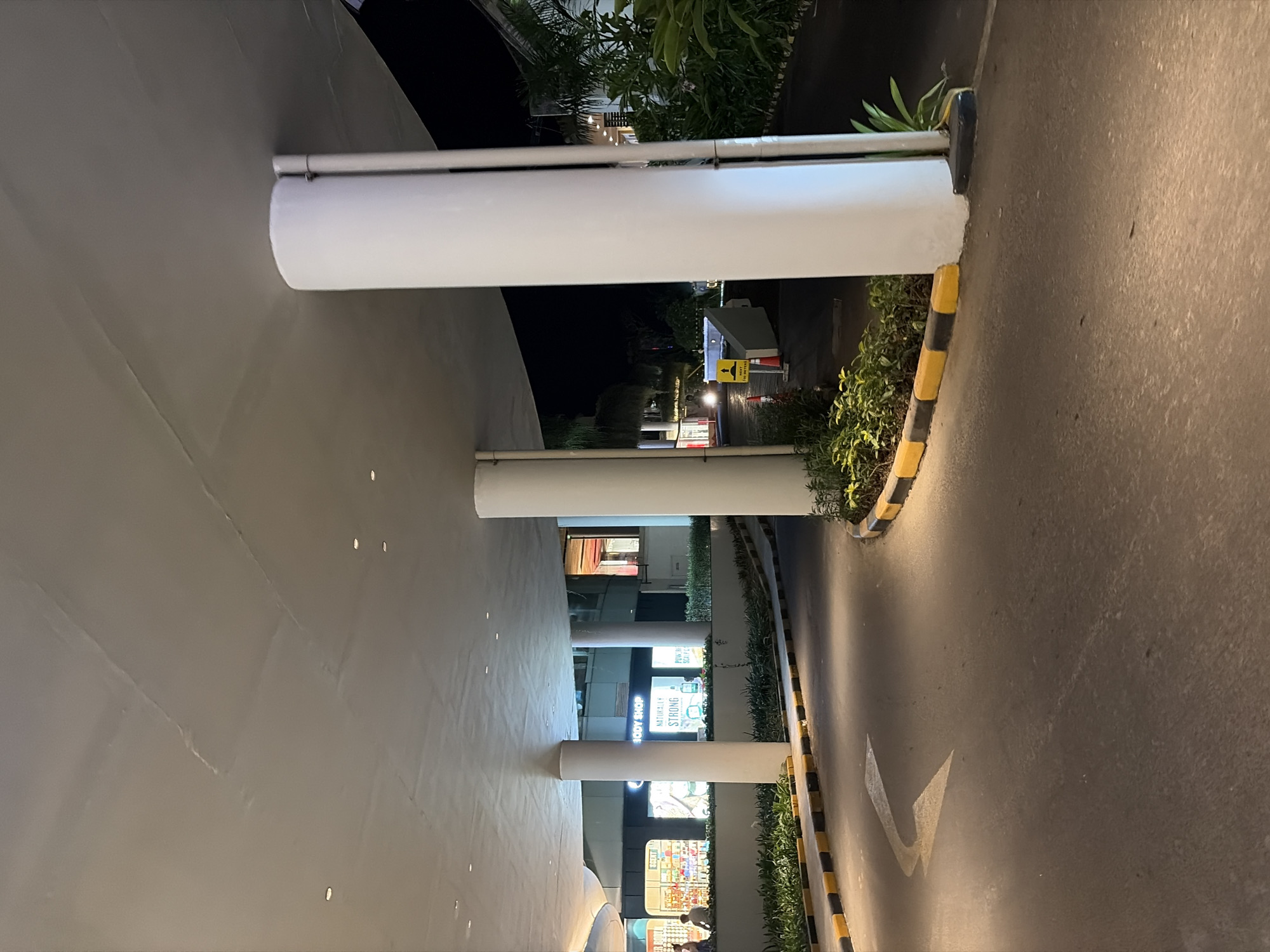 The image captures a view of an overhead structure supported by large white cylindrical pillars, extending over a paved roadway. The roadway has a yellow and black striped curb and a white arrow painted on it, indicating a left turn. To the left of the roadway, there is a landscaped area with green bushes. In the background, through the pillars, a building with illuminated storefronts is visible, with signs for "The Body Shop" and other businesses. There are a few people seated outside one of the establishments. The scene appears to be in the city of Kuta, Indonesia, likely in a commercial or shopping area, and it is nighttime, as indicated by the artificial lighting. The lighting casts shadows and highlights the architectural features of the overhead structure and pillars.