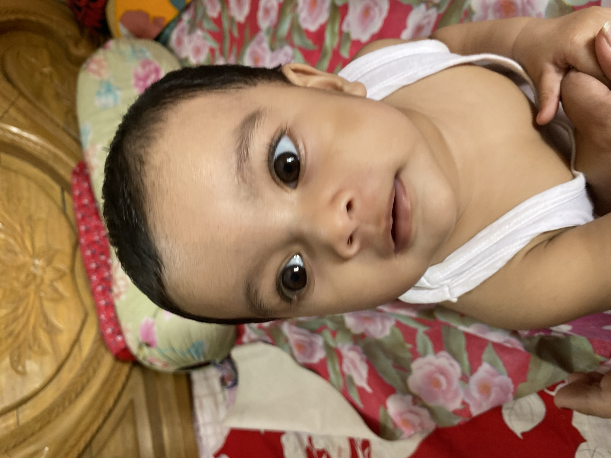 A baby lies on a floral patterned surface. The baby is wearing a white tank top. The surface is located inside and contains a pillow. The location is in Dhaka, Bangladesh.