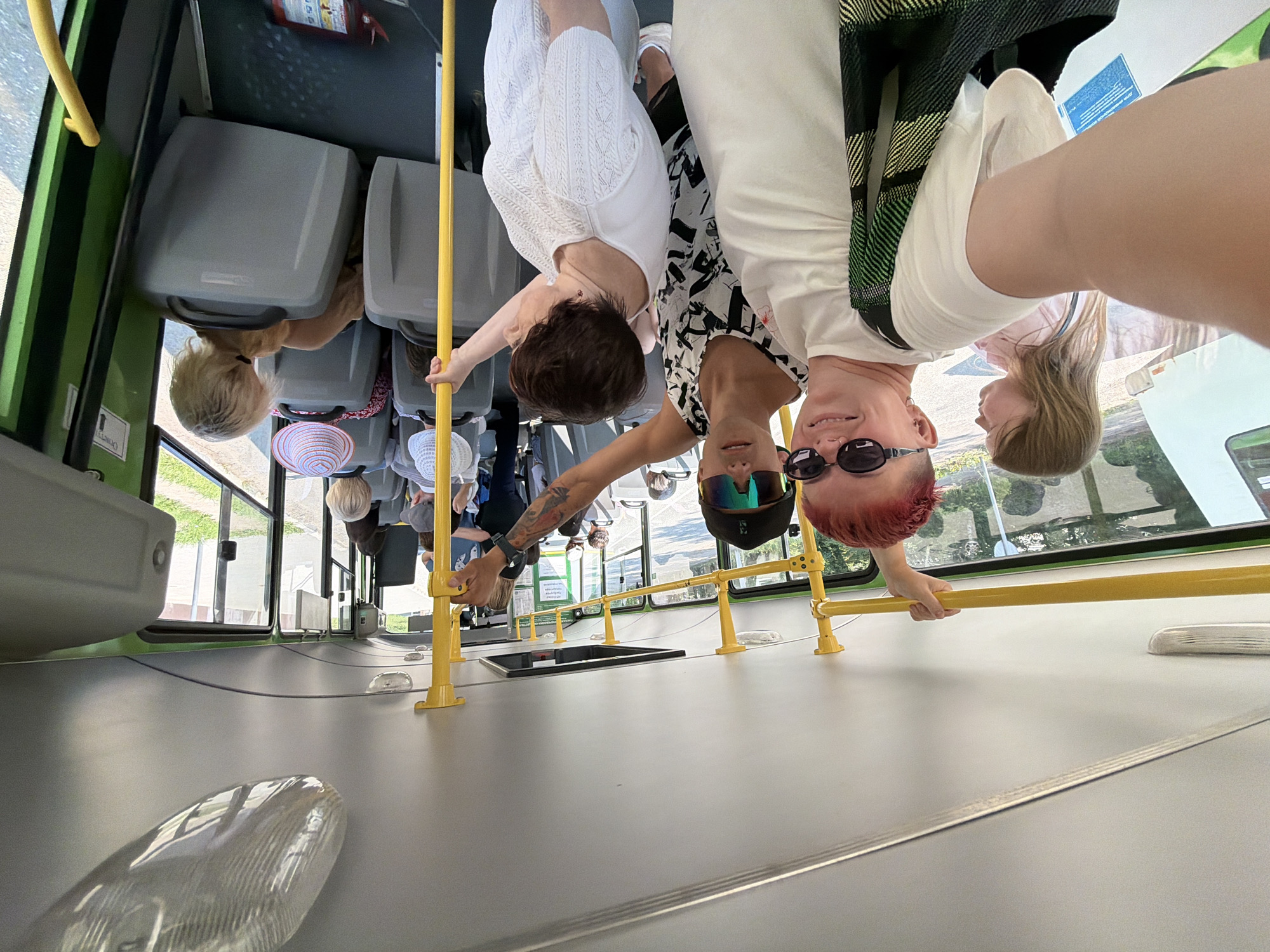 A candid, wide-angle shot captures the interior of a public bus in Chaikovsky, Russia, on a sunny day. Three individuals are prominently featured in the foreground, with others visible in the background. The person on the left, facing the camera and slightly to the right, has bright red hair, sunglasses, and a white t-shirt, carrying a green and black checkered bag. To their right, a second person, also facing the camera, wears sunglasses and a black and white patterned top. A third person, partially visible in the mid-right of the frame, has short brown hair and wears a white top, looking to the right. Several other passengers are seated or standing throughout the bus, some with hats. Yellow poles and handrails are visible throughout the bus interior, typical of public transportation. The bus windows offer a glimpse of a sunlit, outdoor environment with greenery and buildings. The overall scene depicts a casual moment on public transport.