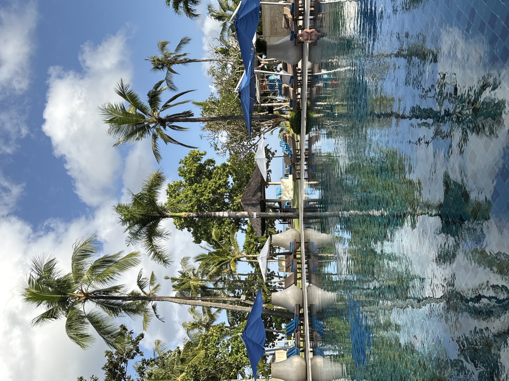 A low-angle shot captures a swimming pool with clear blue water reflecting palm trees and the sky. On the left side of the pool, there are two large gray vase-shaped planters and several blue striped lounge chairs under a large blue umbrella. Further back, a white umbrella shades lounge chairs. To the right of the white umbrella, a thatched-roof gazebo is visible, with lounge chairs and umbrellas beyond it. In the middle ground, a person is partially submerged in the pool on the right side of the frame. Palm trees and lush green foliage surround the pool area, with a bright blue sky dotted with white clouds overhead. This scene appears to be a resort in Kuta Selatan, Indonesia.