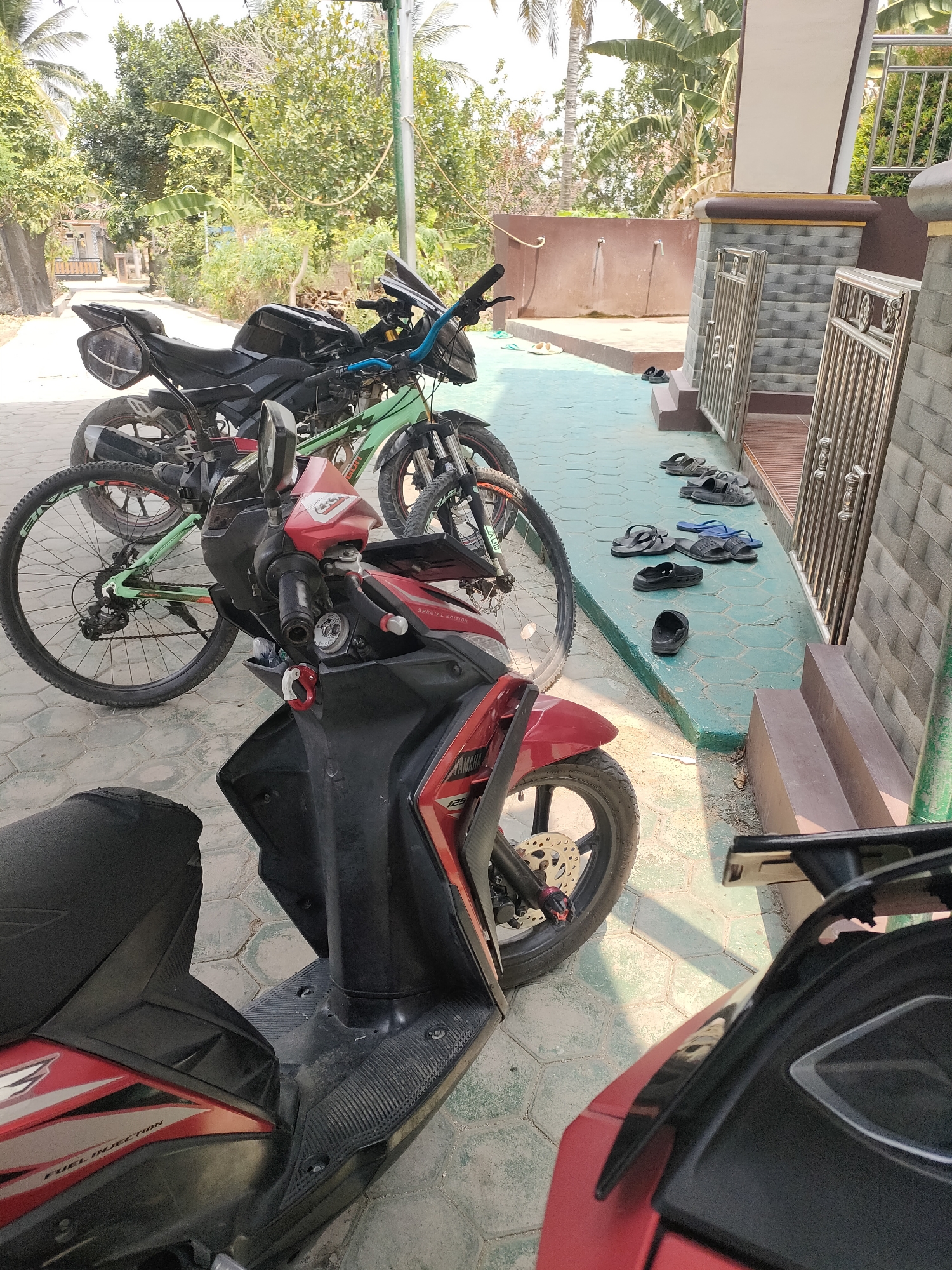 The image depicts an outdoor scene in Pringsewu, Indonesia, likely in front of a residential building. In the foreground, two motorcycles are parked on a patterned concrete floor. The motorcycle on the left is a red scooter, partially visible from the side, showing a "FUEL INJECTION" label. The motorcycle on the right is a black and red scooter, also partially visible. Behind these scooters, a black motorcycle with blue handlebars and a green bicycle are parked. The bicycles' tires and frame are clearly visible. Further in the background, there's a building with a beige and brown facade, a brown wall, and a metal railing. A row of black and blue flip-flops are neatly arranged on a green-painted concrete strip beside the building. Lush green trees and foliage, including palm trees, are visible in the background, suggesting a tropical environment. The lighting indicates it is daytime, with shadows cast on the ground.