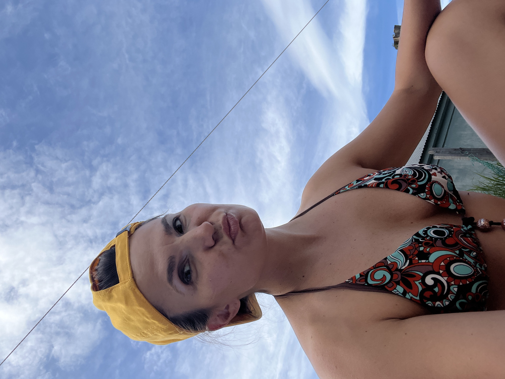 A low-angle, medium shot captures a woman wearing a yellow baseball cap and a patterned bikini against a bright blue sky with wispy clouds. She is looking towards the camera with her lips pursed. The woman's skin is tanned, and her dark hair is pulled back. She is outdoors, with hints of a building and some greenery in the background, suggesting a casual, possibly seaside setting. The image does not contain specific landmarks or visual cues to identify the location as Petropavlovsk-Kamchatsky, Russia.