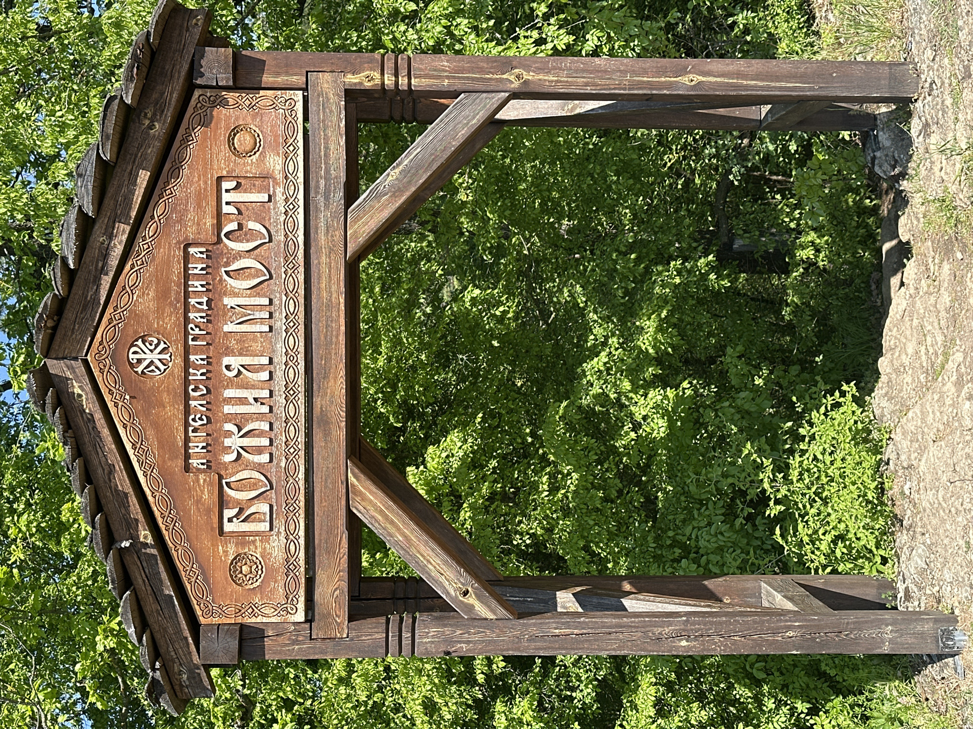 An eye-level, medium shot depicts a weathered, wooden sign in Lilyache, Bulgaria, marking an entrance to the "Angel's Garden – God's Bridge" (Ангелска градина – Божия мост). The sign is mounted on a wooden frame structure composed of vertical posts, a horizontal beam at the top, and angled support beams. The sign itself features Bulgarian text and decorative carvings. The background consists of dense green foliage. The ground at the base of the structure is covered in stone and grass.