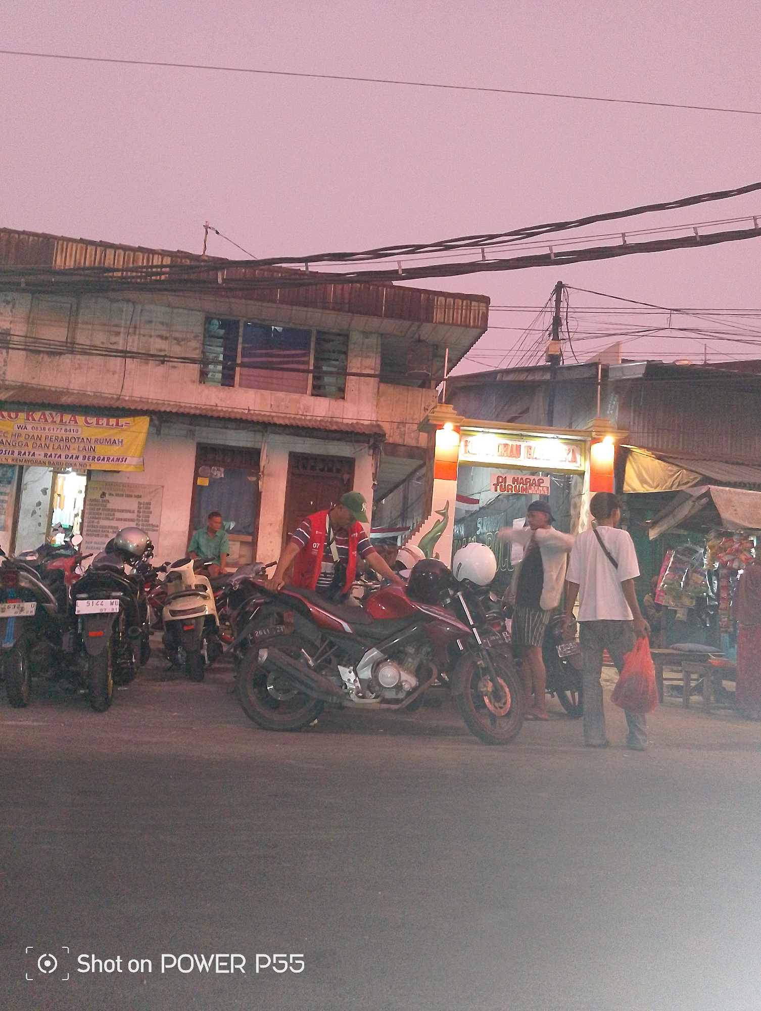 In Surabaya, Indonesia, at dusk, several motorbikes are parked along a street in front of a row of shops. A man in a red vest stands near a red motorbike. Another man, wearing a green shirt, sits on a motorbike. Two other individuals are visible in the background near shop entrances, one holding a bag. The street is paved, and overhead power lines crisscross the scene, illuminated by streetlights from the shops. The sky is a soft pink, indicating the time of day.